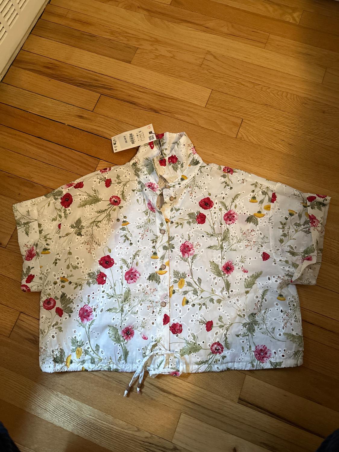 White blouse with floral print in red, green, and yellow tones, on a wooden floor. Visible tag on the collar.