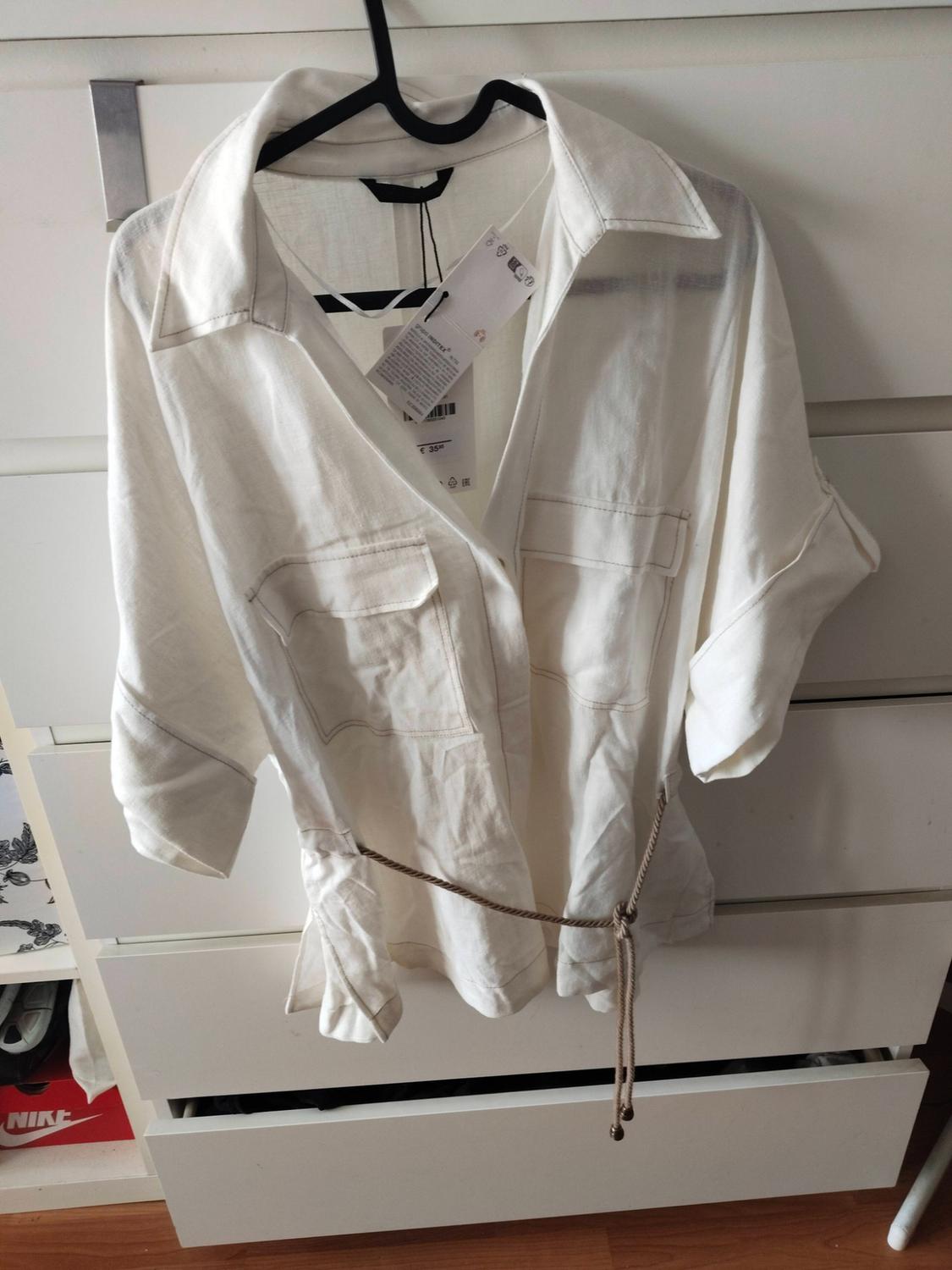 White short-sleeved shirt with a collar, pockets, and rope belt, hanging in front of a white chest of drawers. Below, a red box with NIKE text.