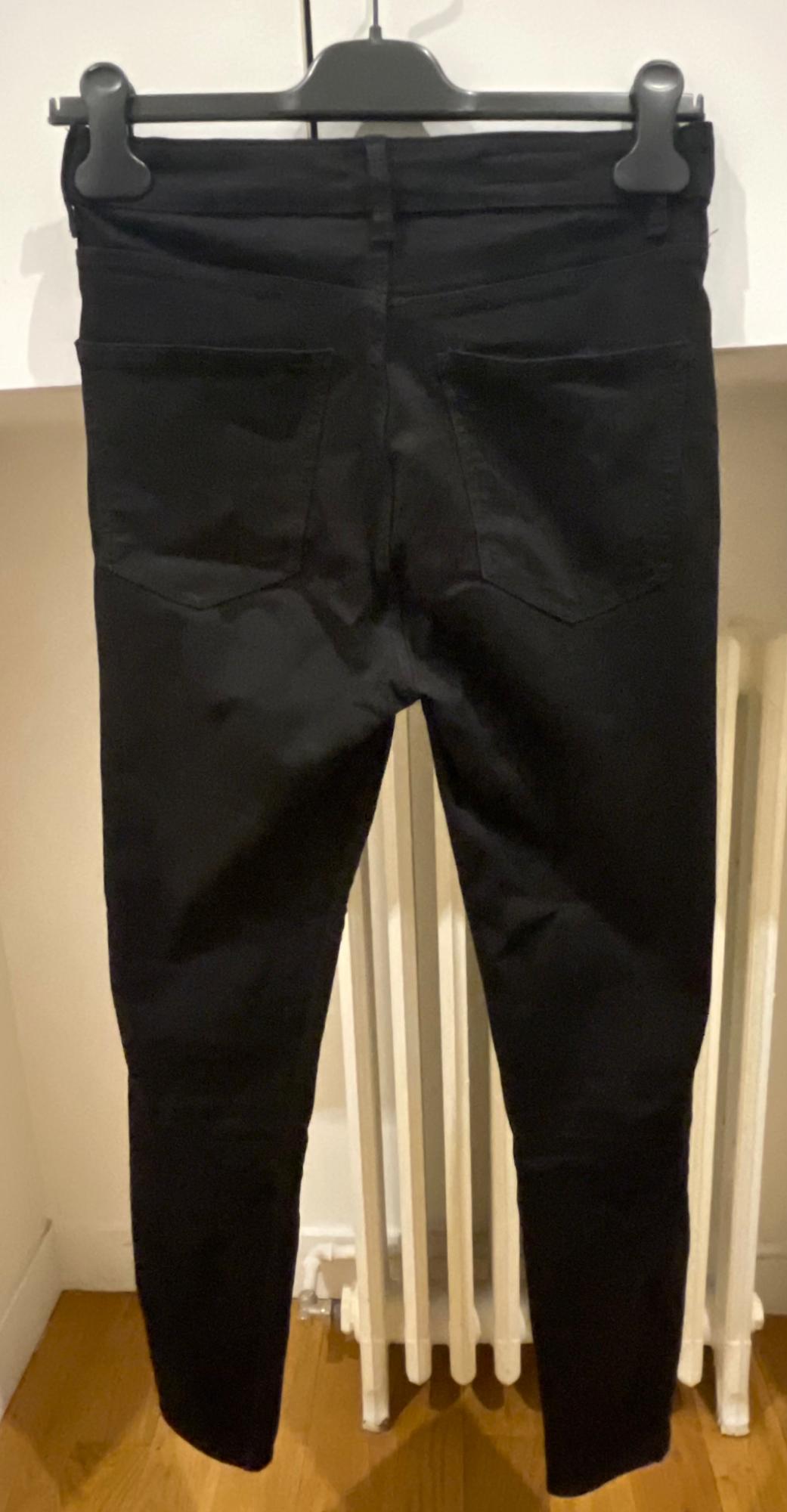 Black trousers hanging on a hanger, viewed from behind. Rear pockets are visible, along with a white wall background and wooden floor.