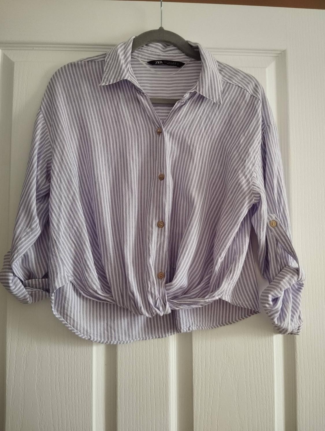 White and lilac striped Zara shirt hanging on a hanger on a white door. Rolled-up sleeves and knot detail at the hem.