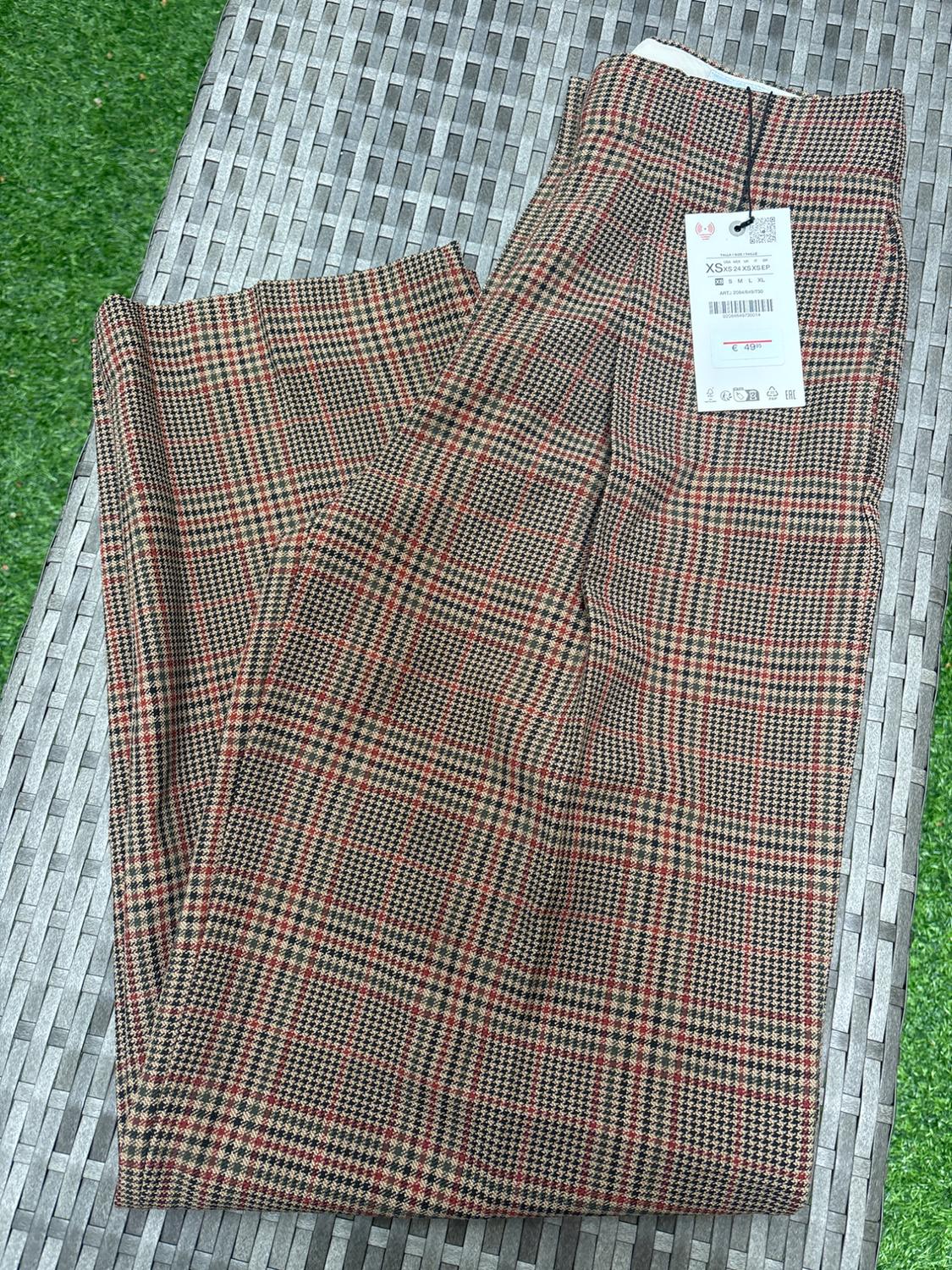 Brown and red checked trousers with a price tag on a grey wicker surface, green grass background.
