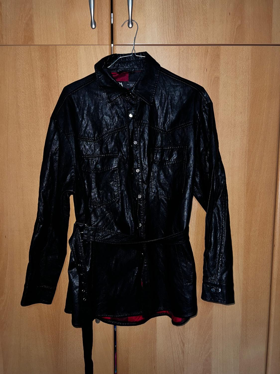 Black leather shirt hanging on a hanger in front of a wooden wardrobe. Detail of buttons and matching belt.