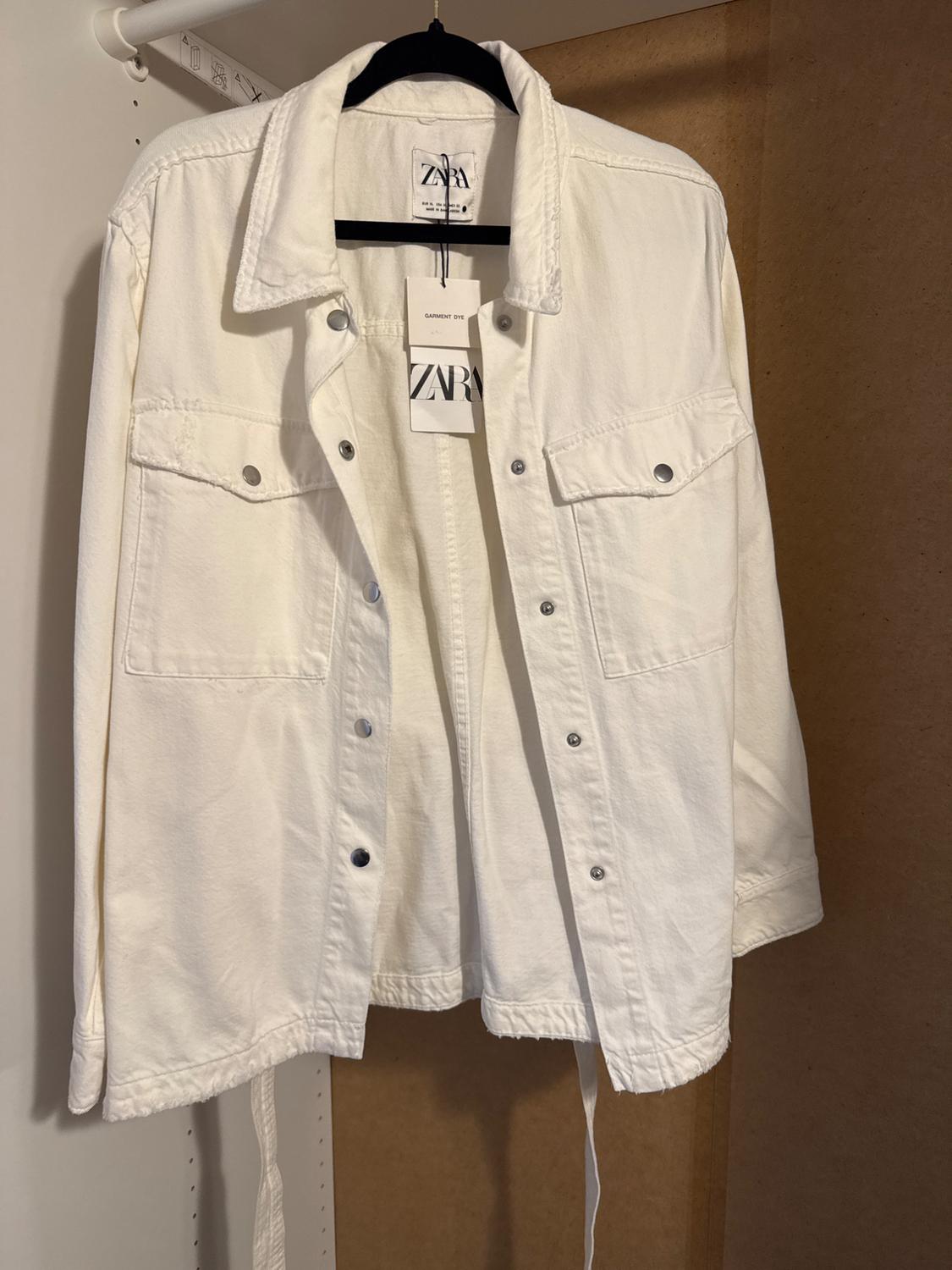 White jacket hanging on a black hanger, with metallic buttons and front pockets. Visible label with the text "ZARA".