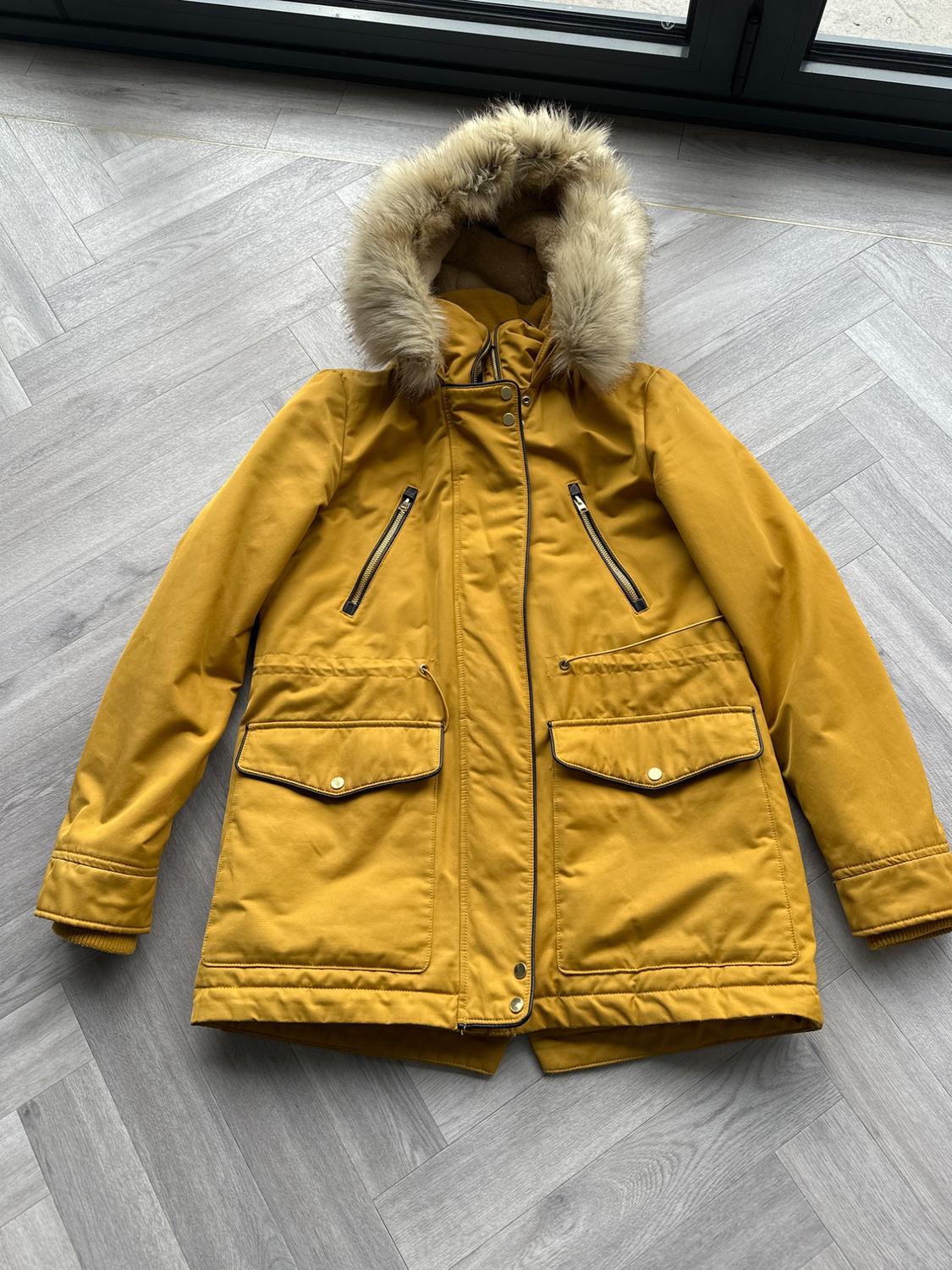 Yellow coat with synthetic fur hood, flap pockets and zips, on grey wooden floor.