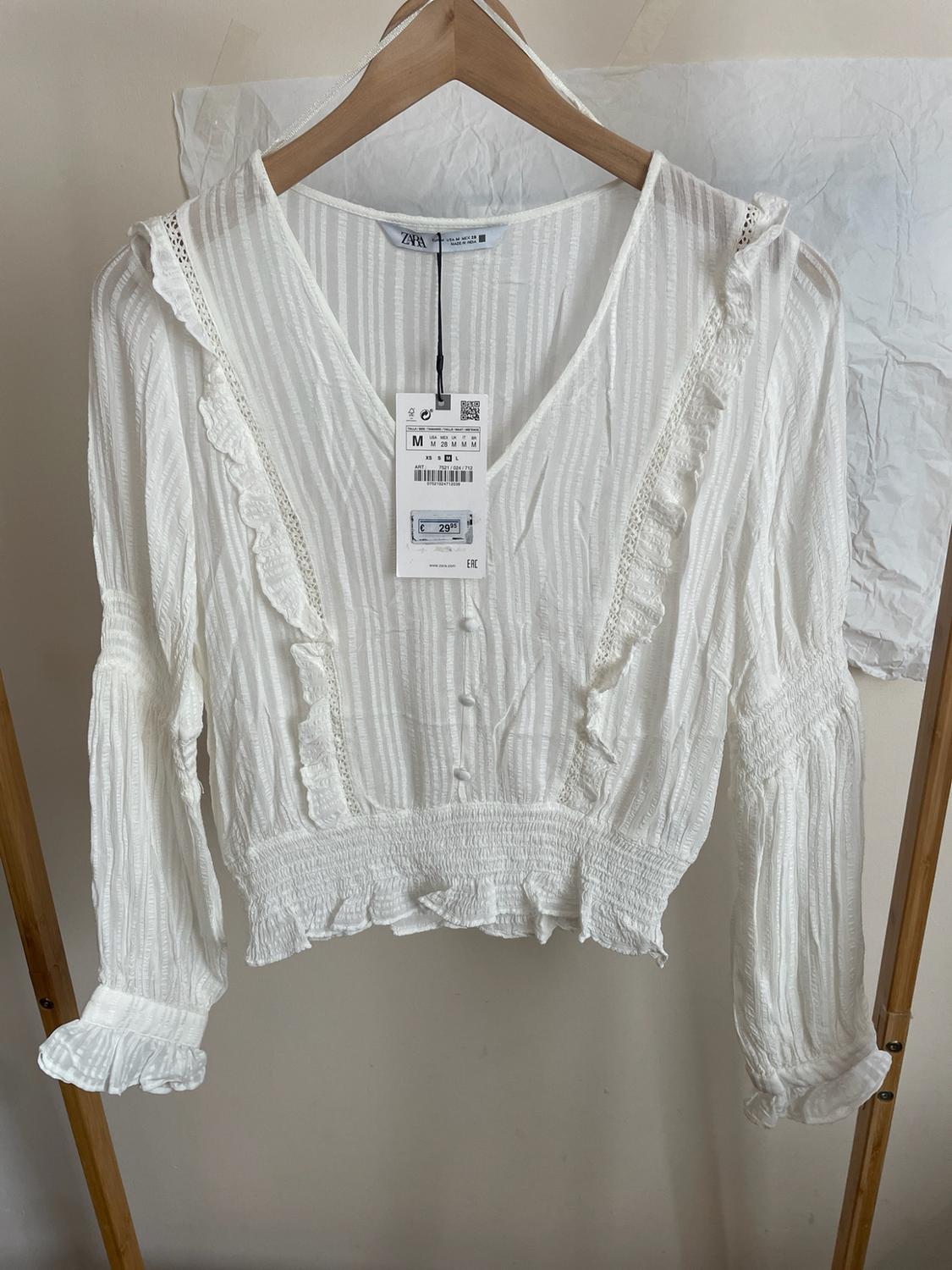 White long-sleeved frilled blouse with buttons and an elasticated waist, hanging on a wooden hanger. Label with text ZARA M €29.95.