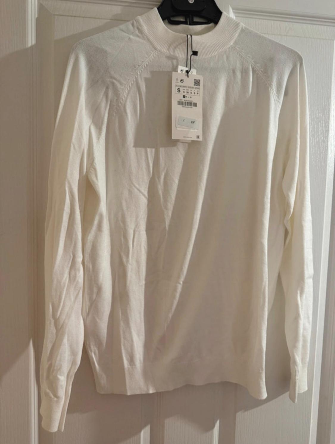 White, long-sleeved jumper hanging on a black hanger, with a visible label showing size S and a barcode.