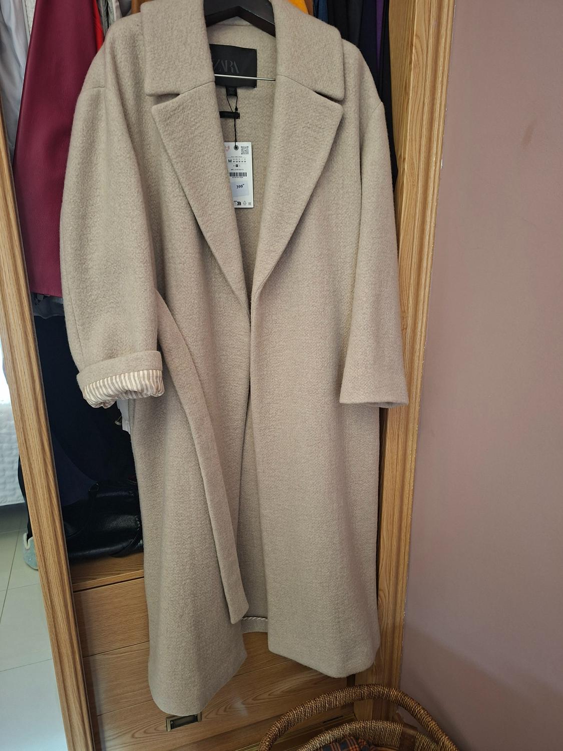 Beige coat hanging in a wooden wardrobe, with a visible label. Background with colourful clothes and a basket at the bottom.