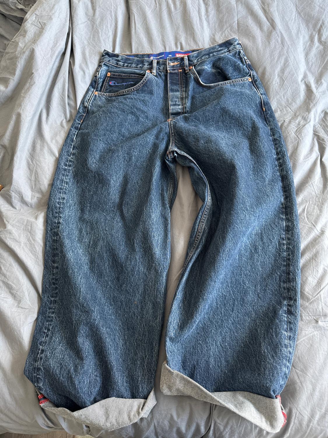 Blue, wide-leg jeans with a hem, placed on a grey bed. Visible logo pocket.