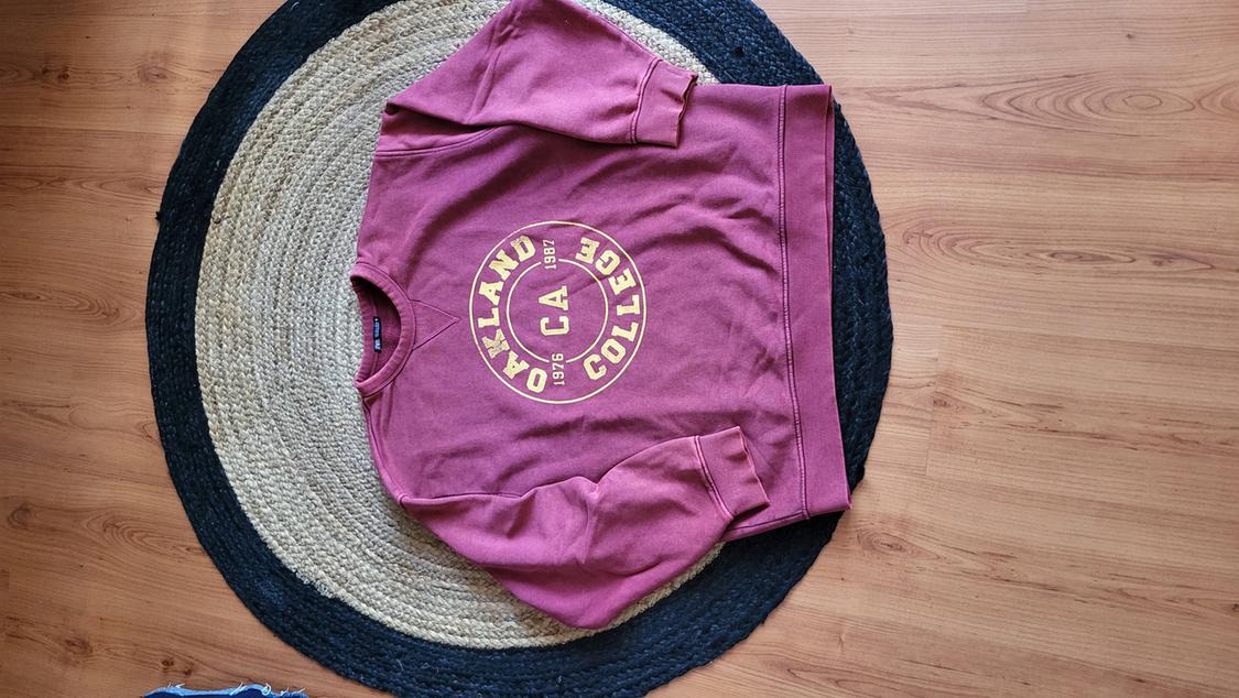 Burgundy sweatshirt with "Oakland College CA 1976 1981" text in yellow, on a round rug on a wooden floor.