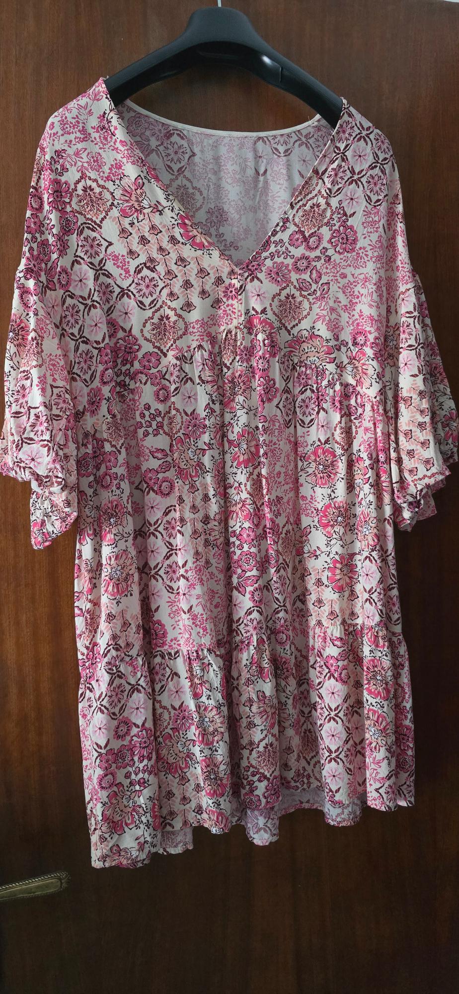 Short-sleeved blouse with a floral print in pink and white tones, hanging on a black hanger against a wooden background.