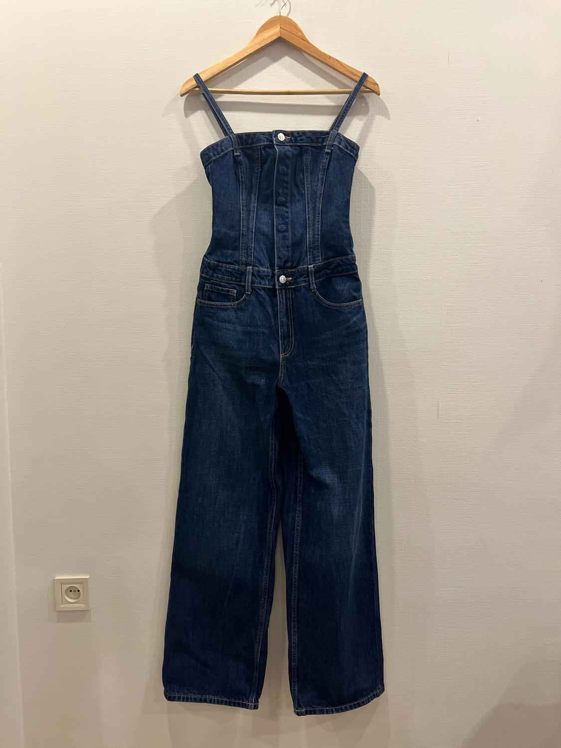 Dark blue denim dress with thin suspenders and a front button placket, hanging on a wooden hanger on a light wall.