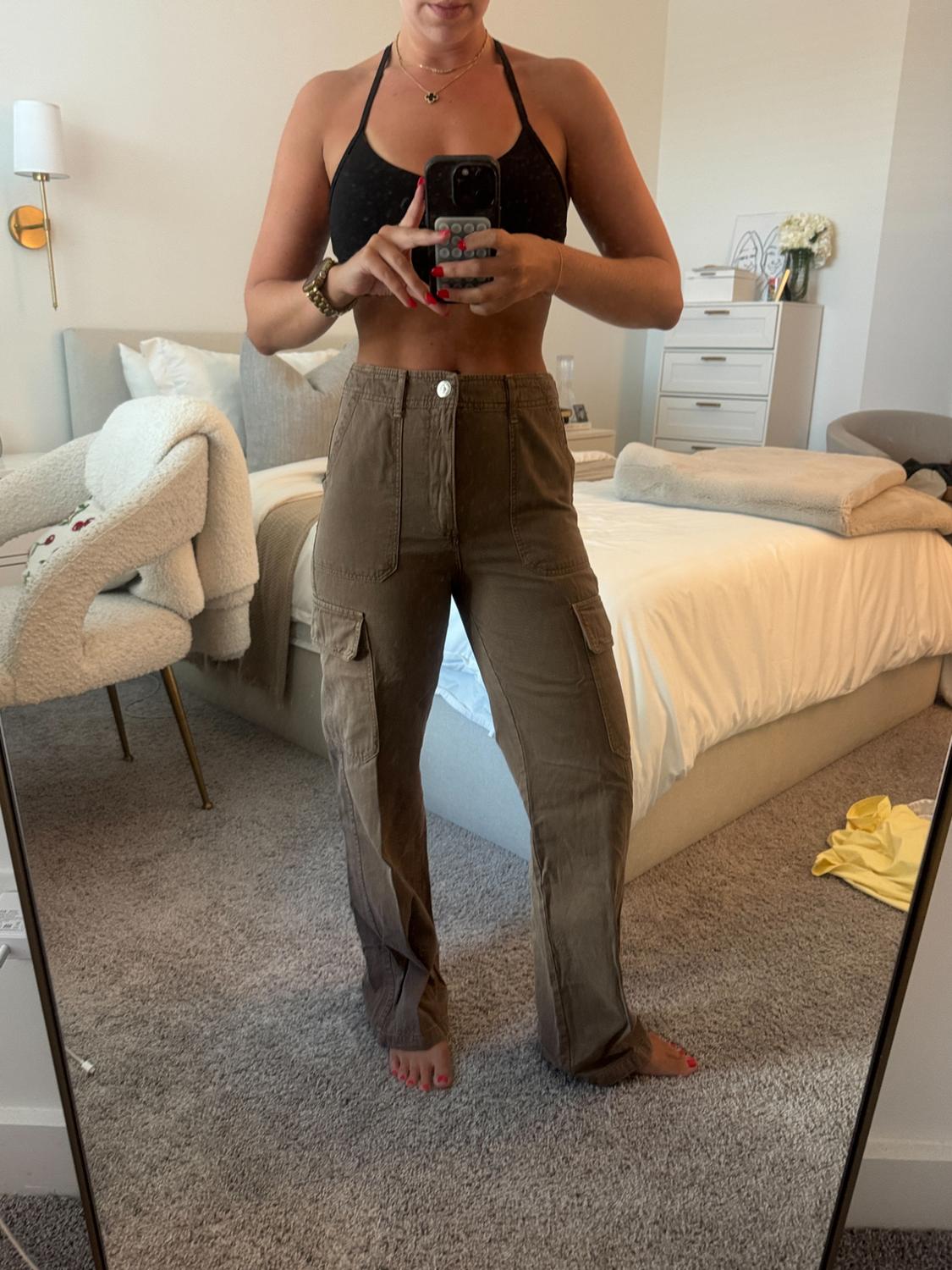 Woman taking a picture in front of the mirror in her bedroom. She's wearing a black top and brown cargo trousers. Relaxed and casual atmosphere.