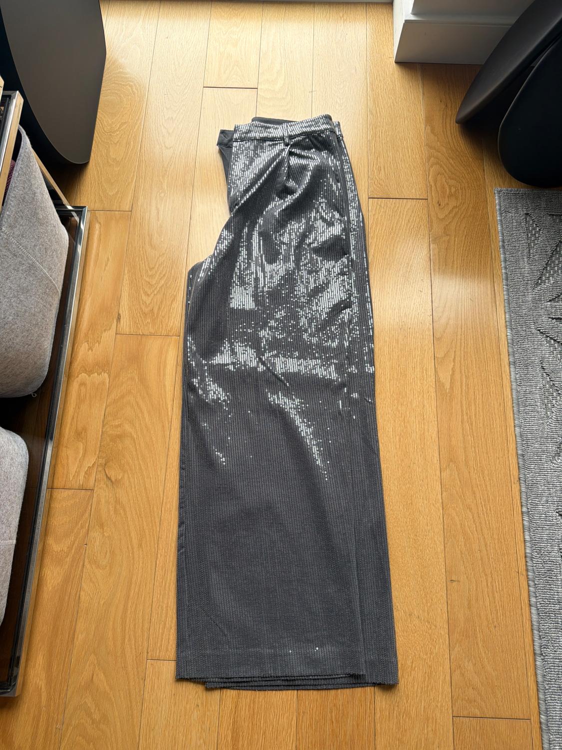 Shiny gray pants on a light wood floor, next to dark furniture and a gray rug. Natural light from the window.