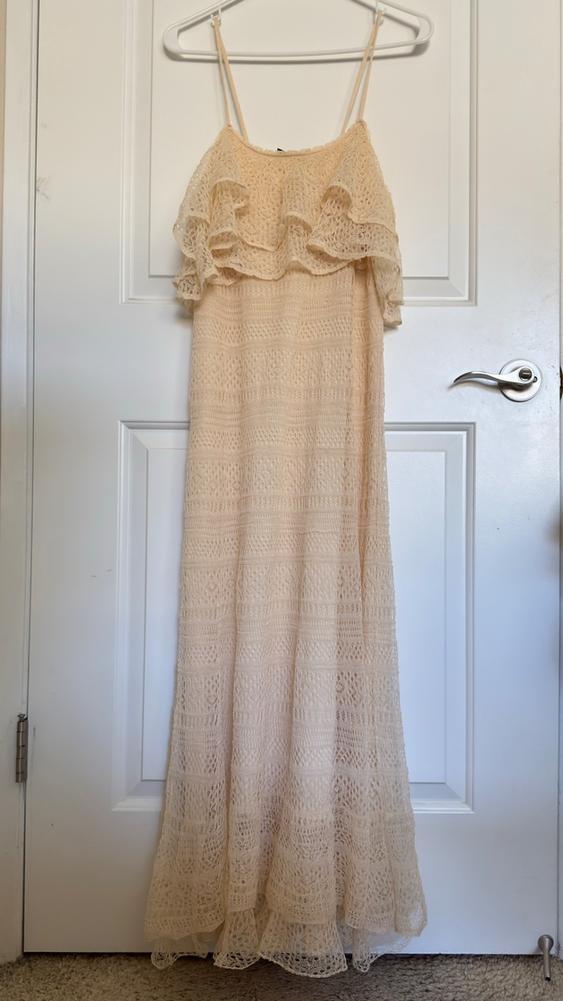 Long beige lace dress with spaghetti straps and ruffles on the chest, hanging on a white hanger over a white door.