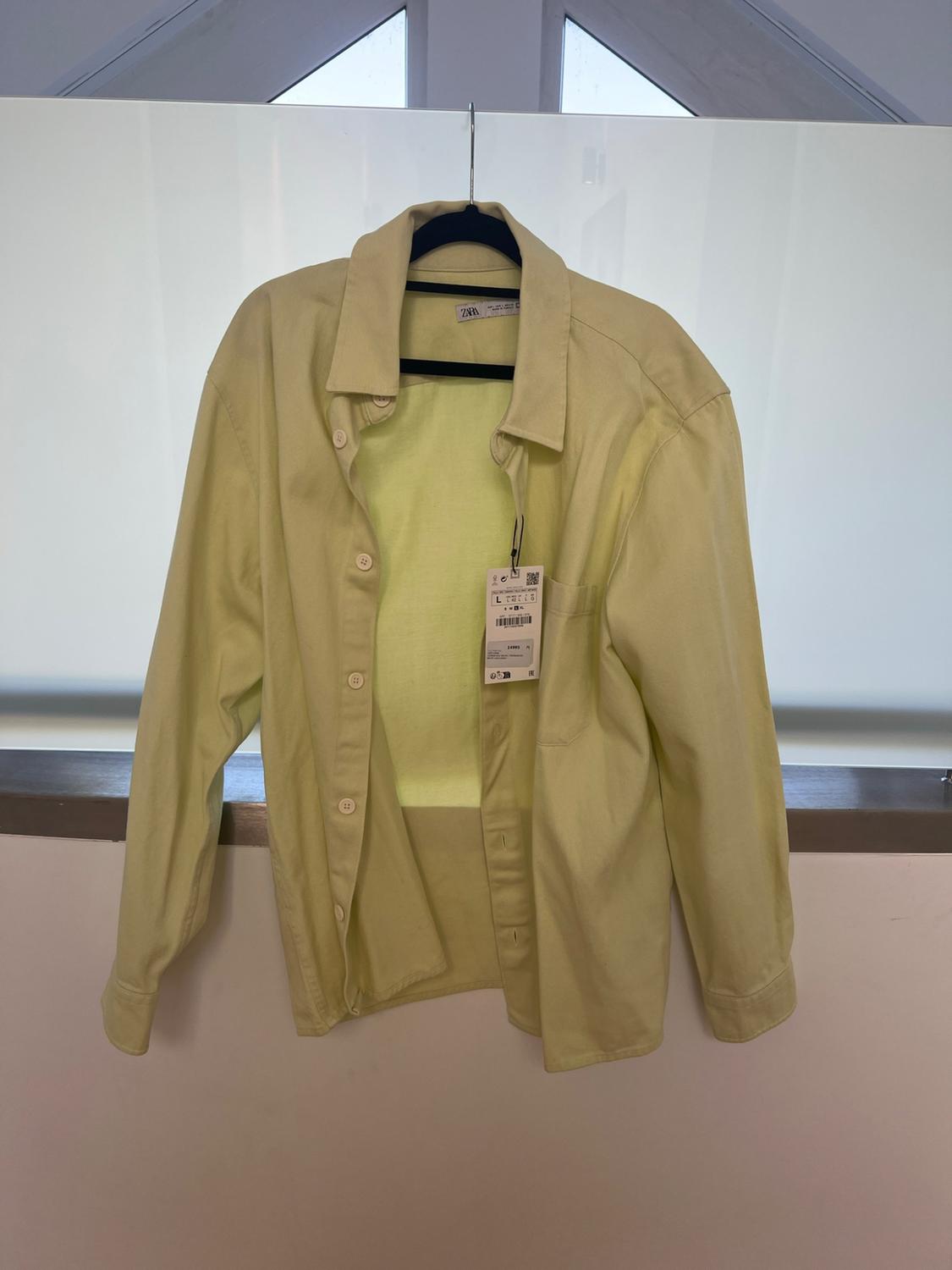 Yellow long-sleeved unbuttoned shirt on a black hanger in front of a window. ZARA can be read on the neck label.
