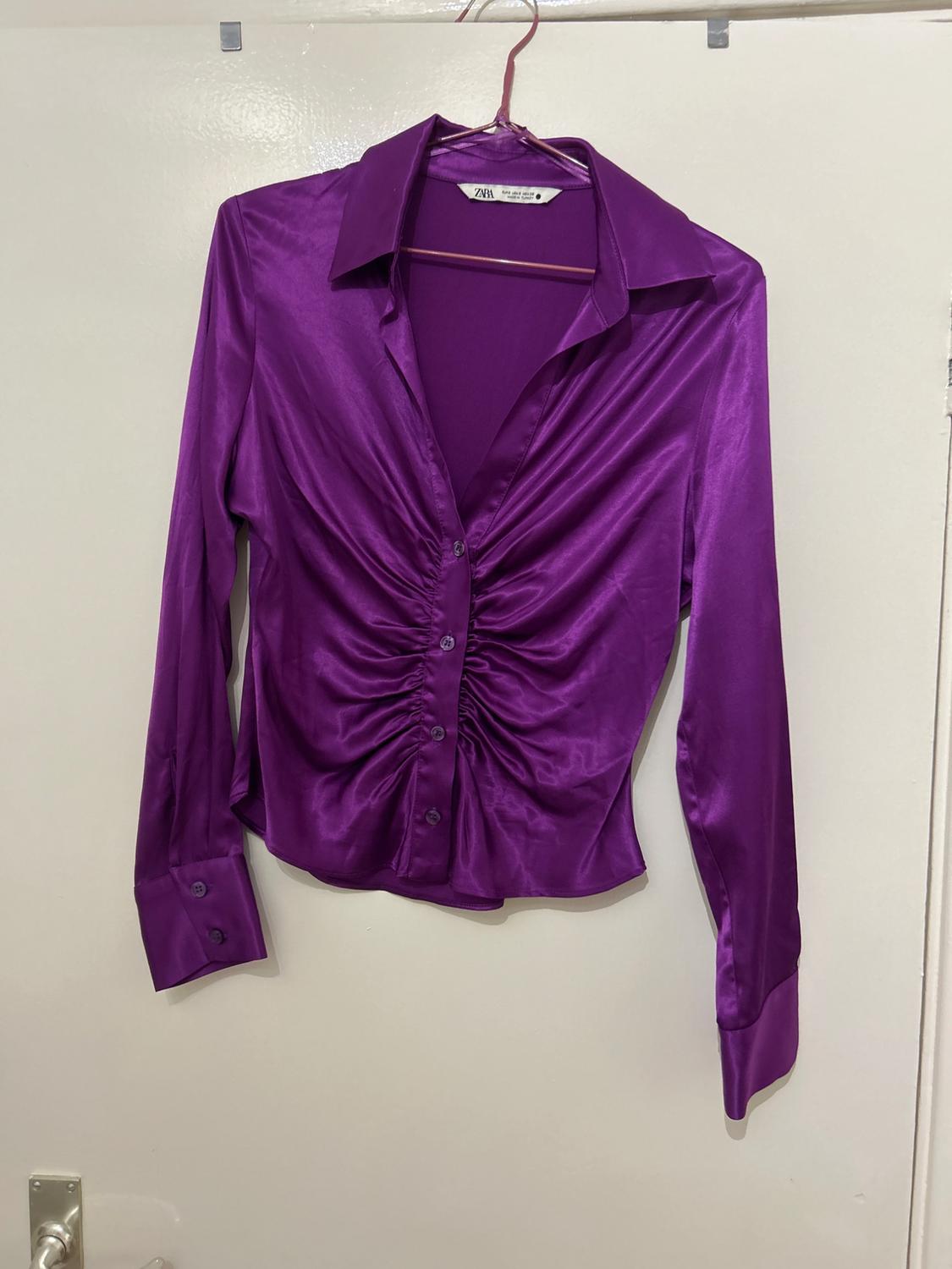 Purple satin blouse with ruching on the front and a classic collar, hanging on a hanger. Visible label: "Zara".