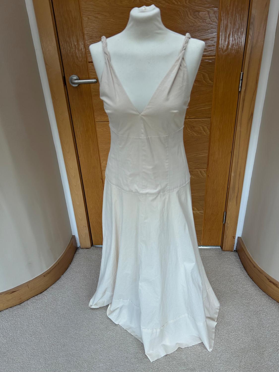 Cream-coloured maxi dress with a V-neck and braided straps on a mannequin, in front of a wooden door in an indoor setting.