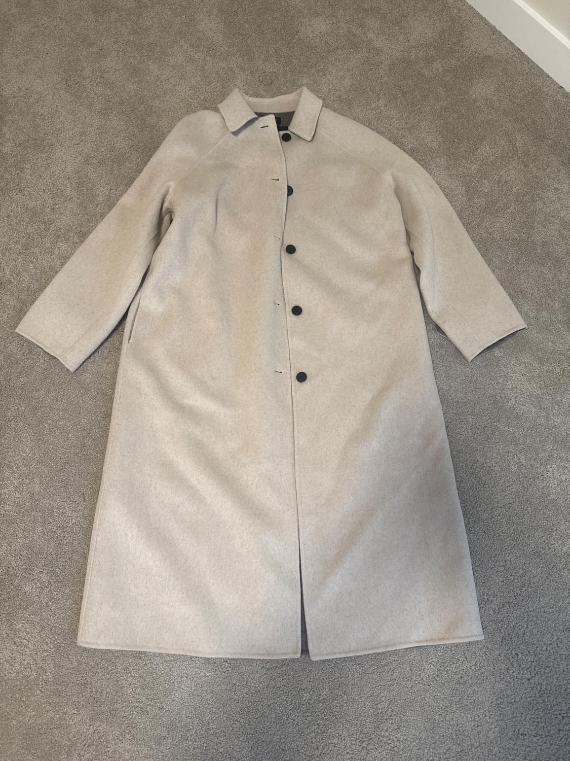 Beige wool coat with black buttons, long-sleeved, spread out on a gray rug.