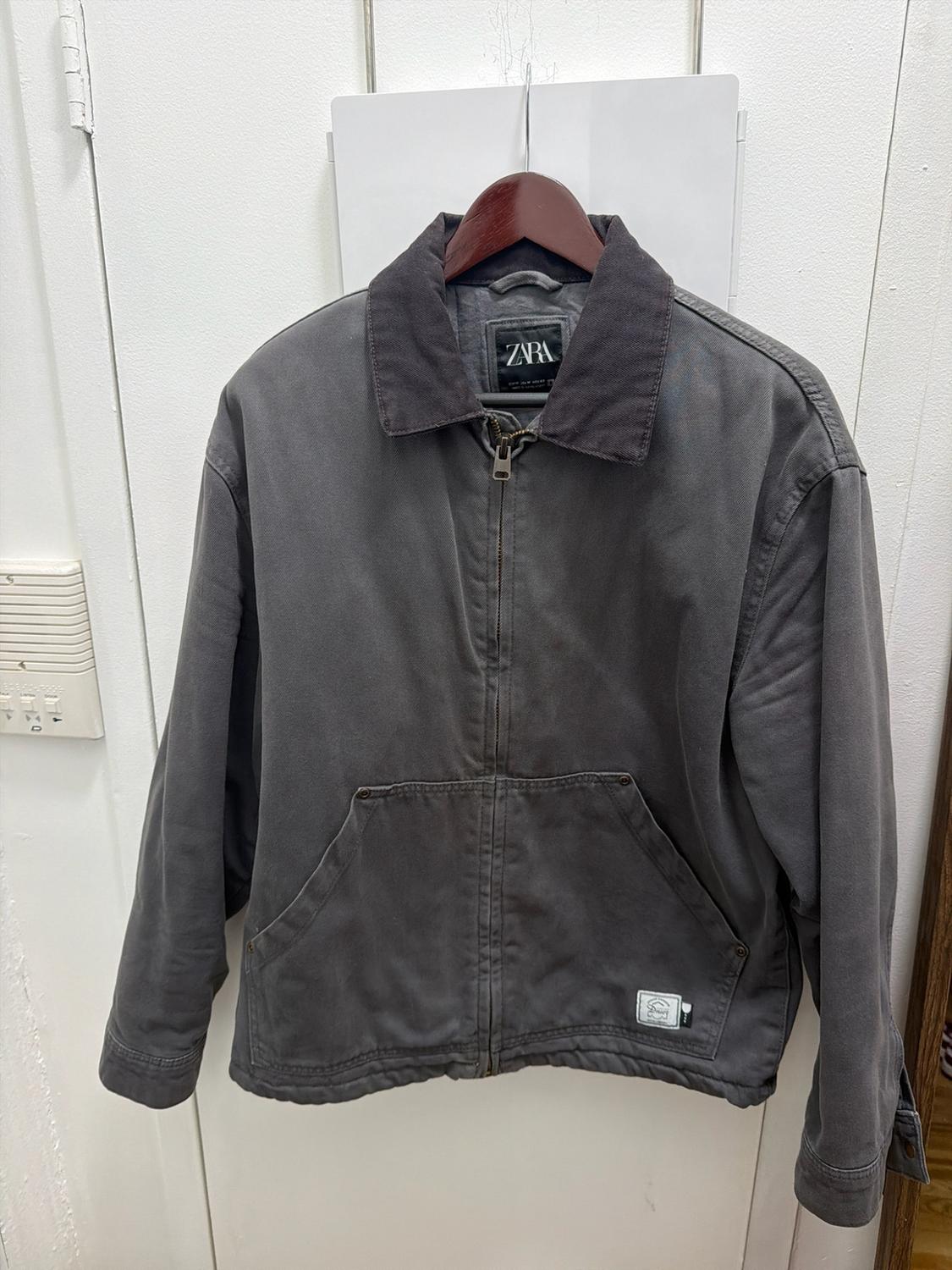 Dark gray jacket with a front zipper and contrasting collar hanging on a white door. Inner label with ZARA text.