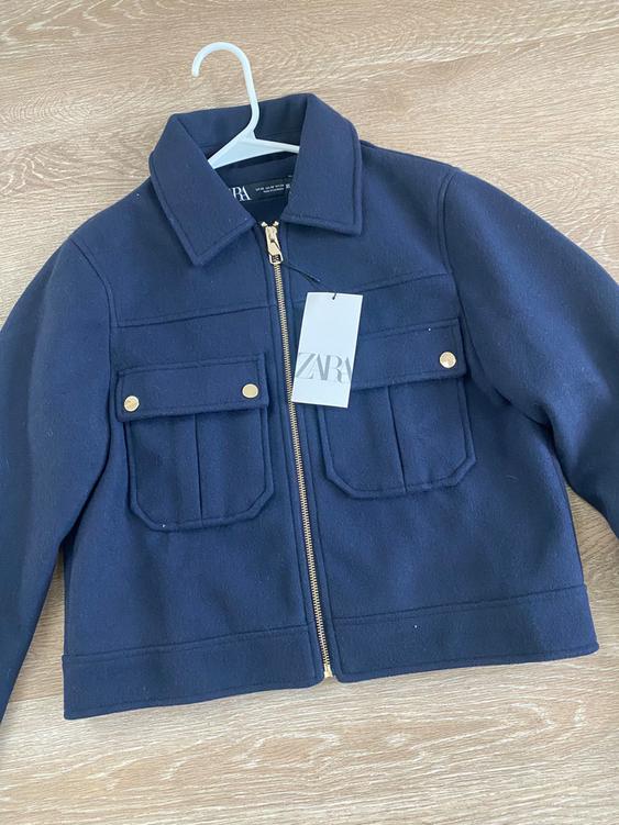 Navy blue jacket with a gold zipper and two front pockets, hanging on a white hanger. Visible tag with text "ZARA".