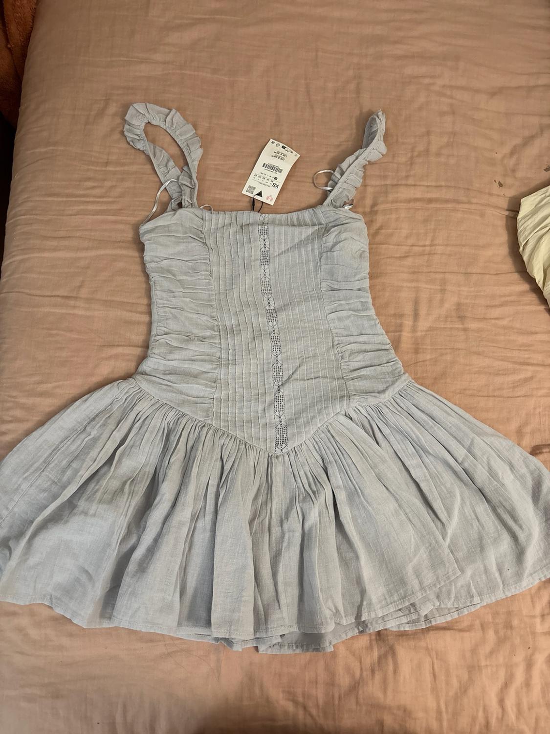 Short blue-grey dress with ruffled straps, shirred bodice, central lace trim and pleated skirt, on a bedsheet.