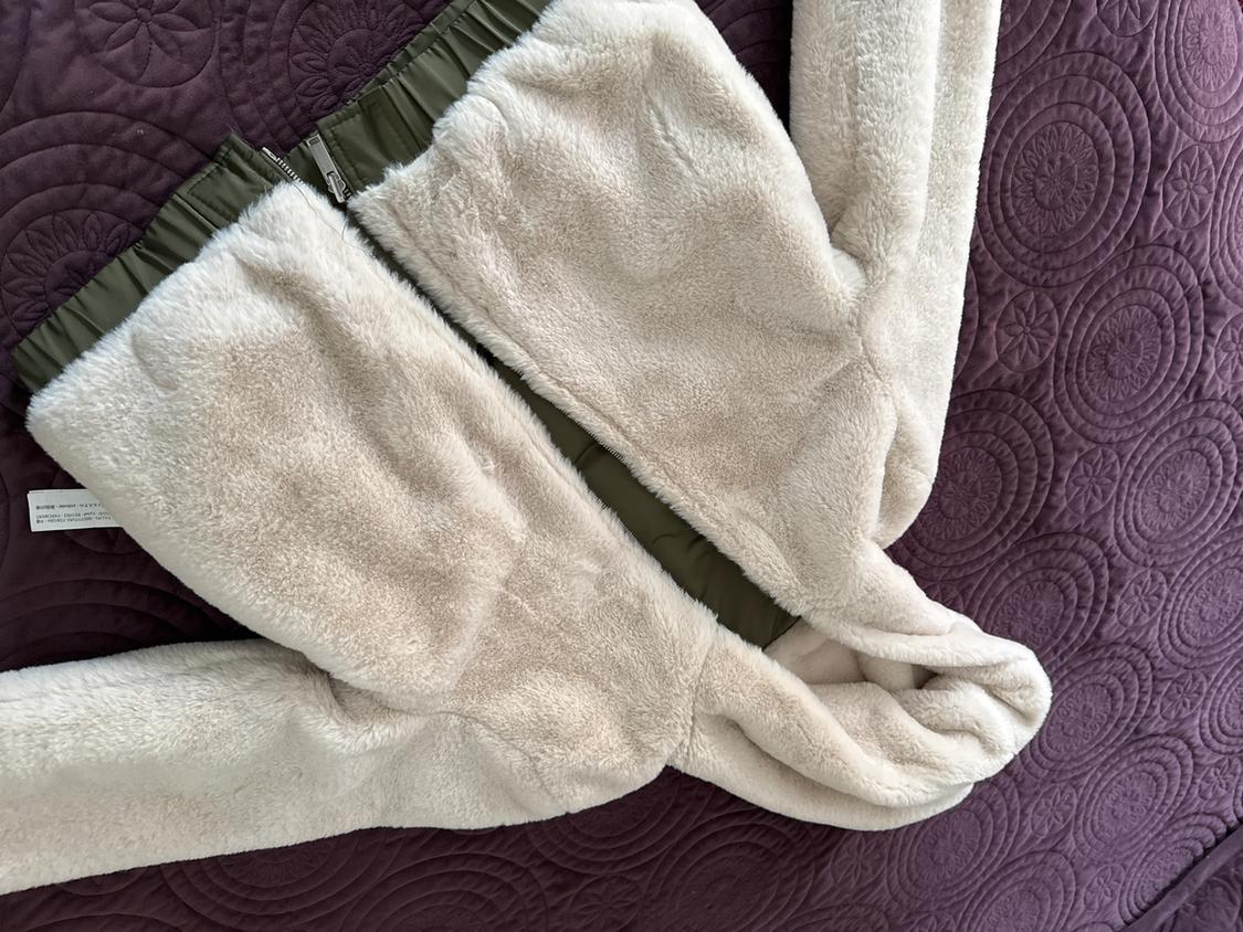 Beige fleece jacket with a hood and dark green details, on a purple bedspread with an embossed circle design.