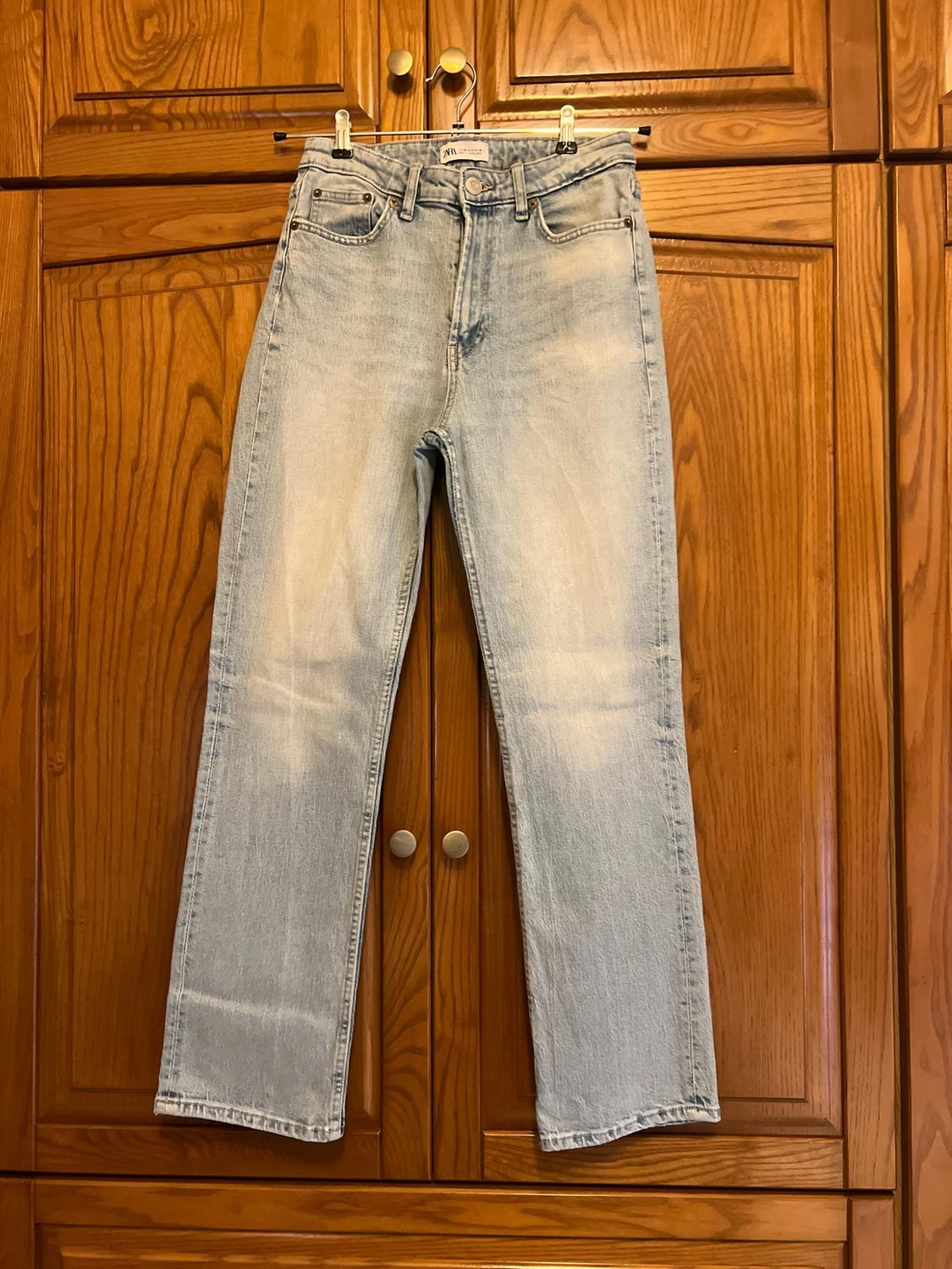 Light blue straight-leg washed jeans, hanging on a metal hanger in front of a wooden wardrobe. Casual style.