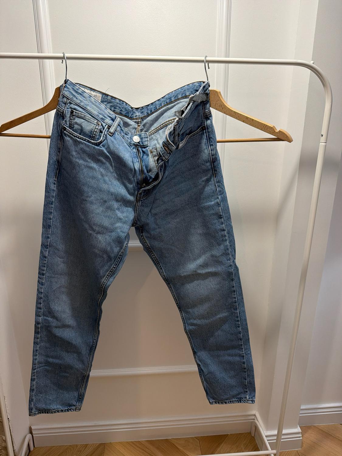 Unbuttoned blue jeans, hanging on a wooden hanger on a white coat rack. White wall background and wooden floor.