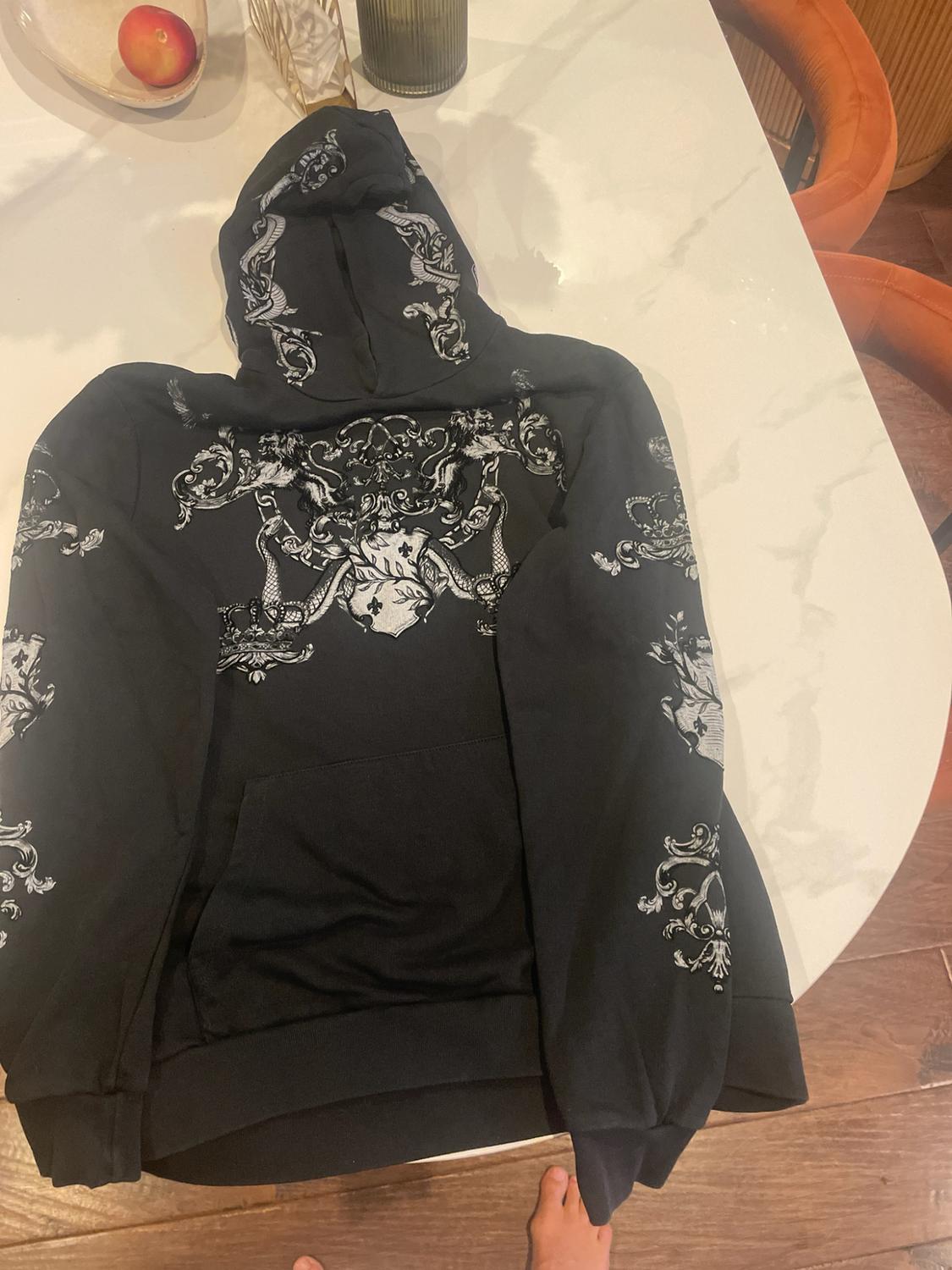 Black sweatshirt with a white ornamental print on a white table. Relaxed atmosphere, with wooden flooring and bare feet visible.