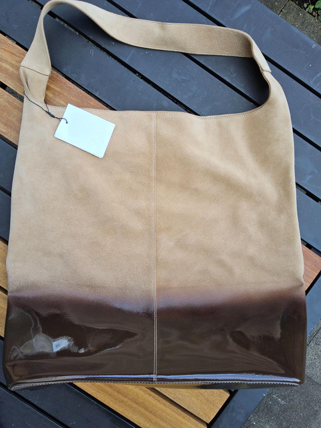 Beige shoulder bag with a shiny brown base, on a wooden and metal table. White tag hanging from the handle.