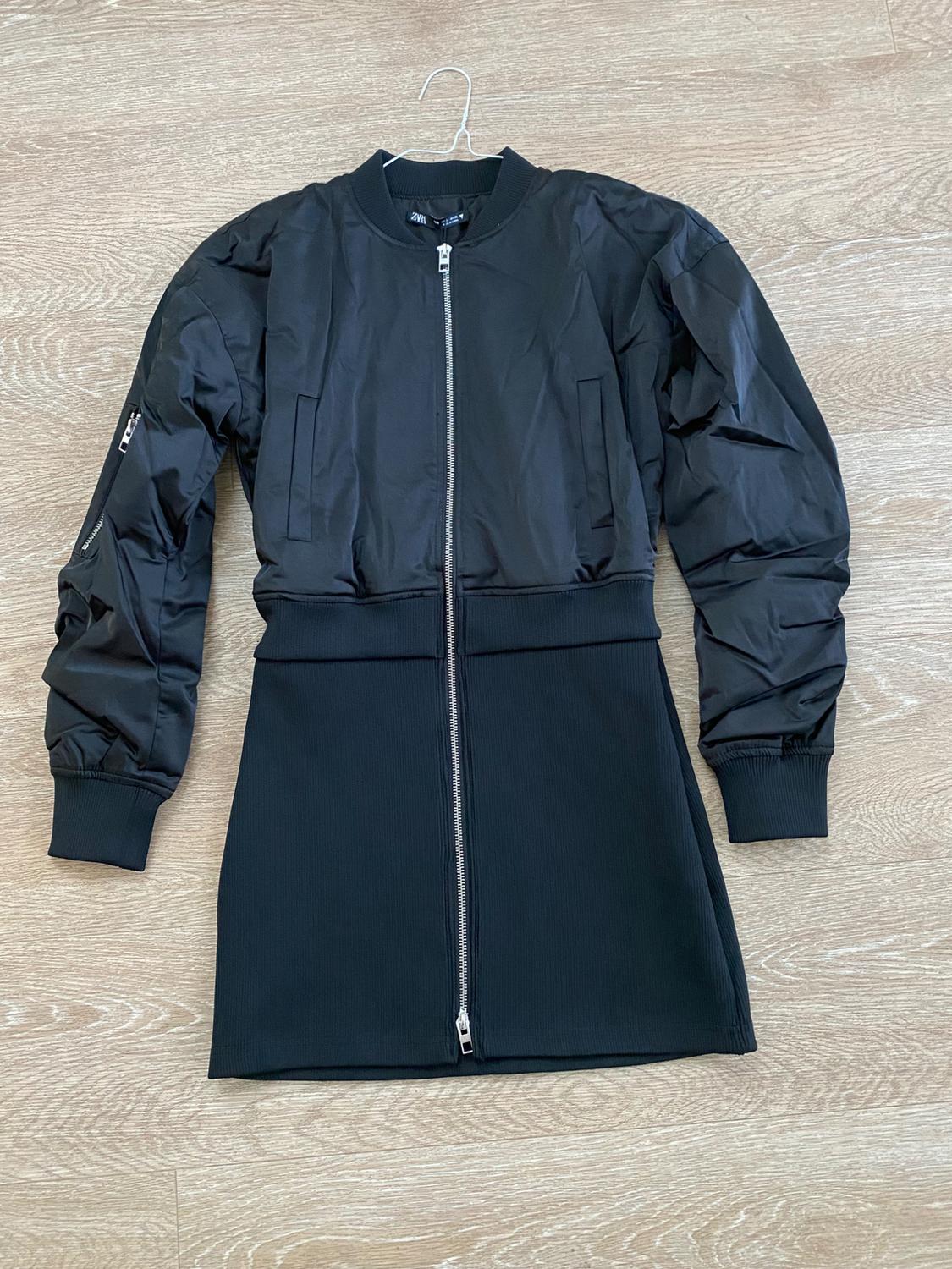 Black dress with a front zipper and long bomber sleeves, hanging on a hanger over a light wood floor.