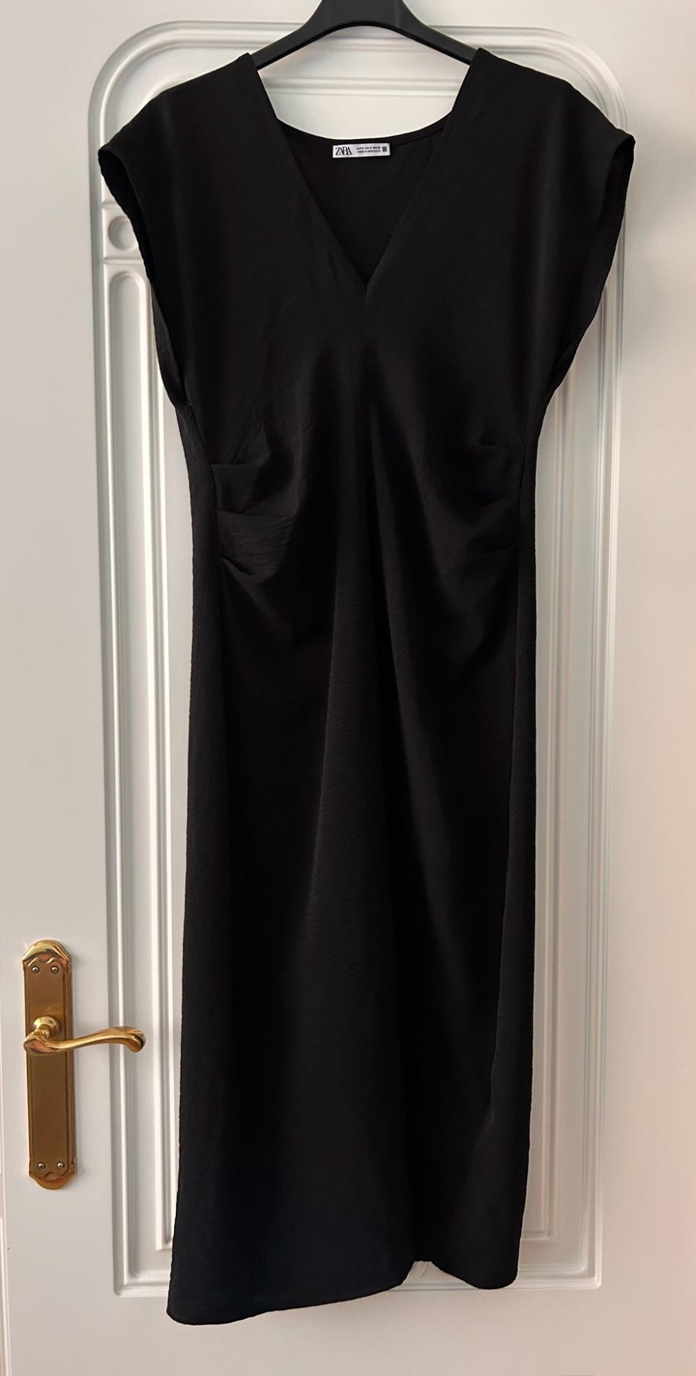 Black V-neck short-sleeved dress with gathering, hanging on a white door with a gold handle. Text ZARA.