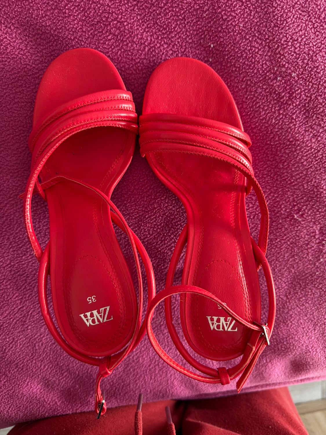 Red strappy sandals with buckle on a pink textured background. Visible text: "ZARA 35".