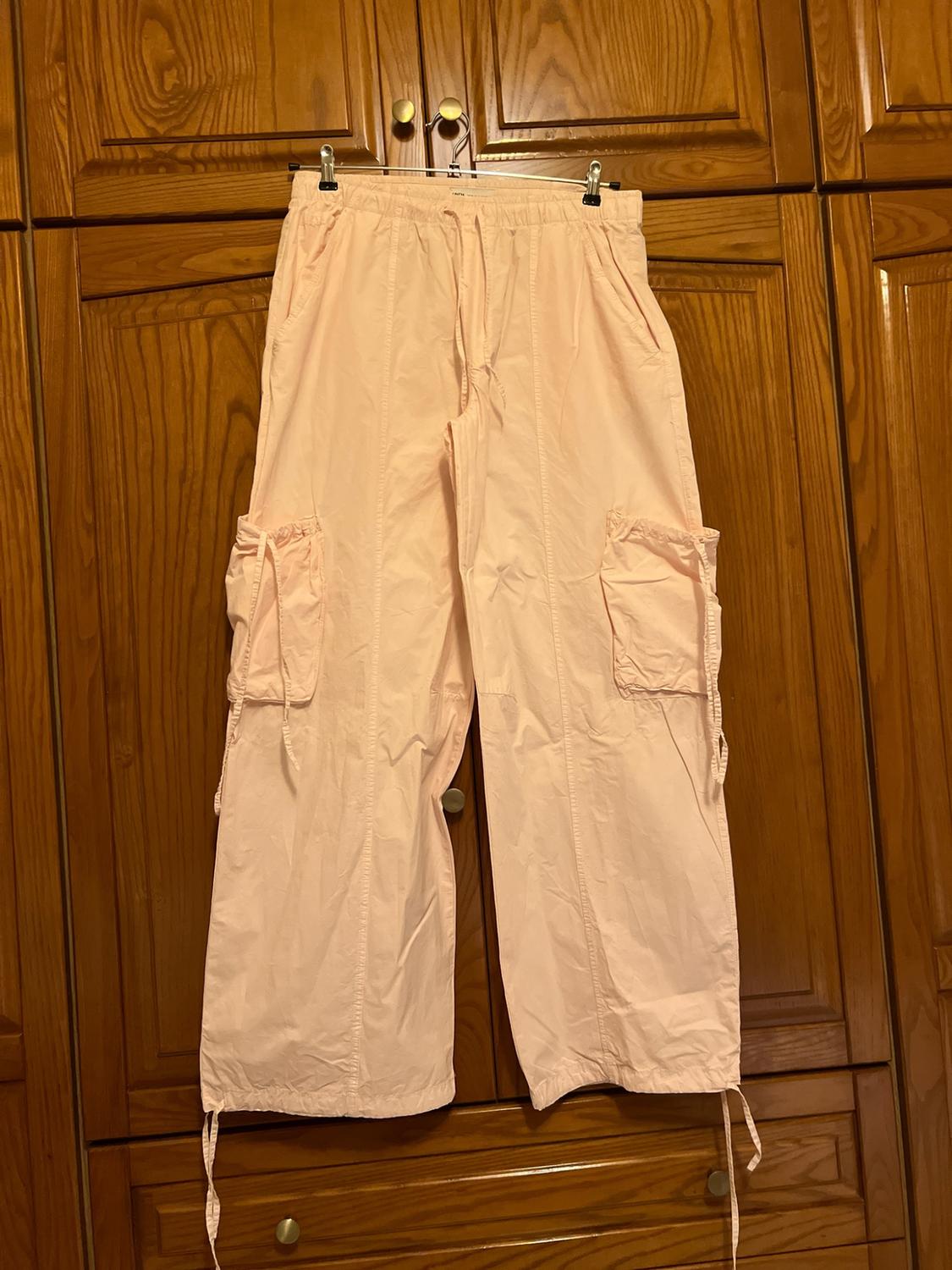 Light pink cargo trousers hanging in a wooden wardrobe. Casual style with an elasticated waistband, side pockets and drawstrings at the hems.