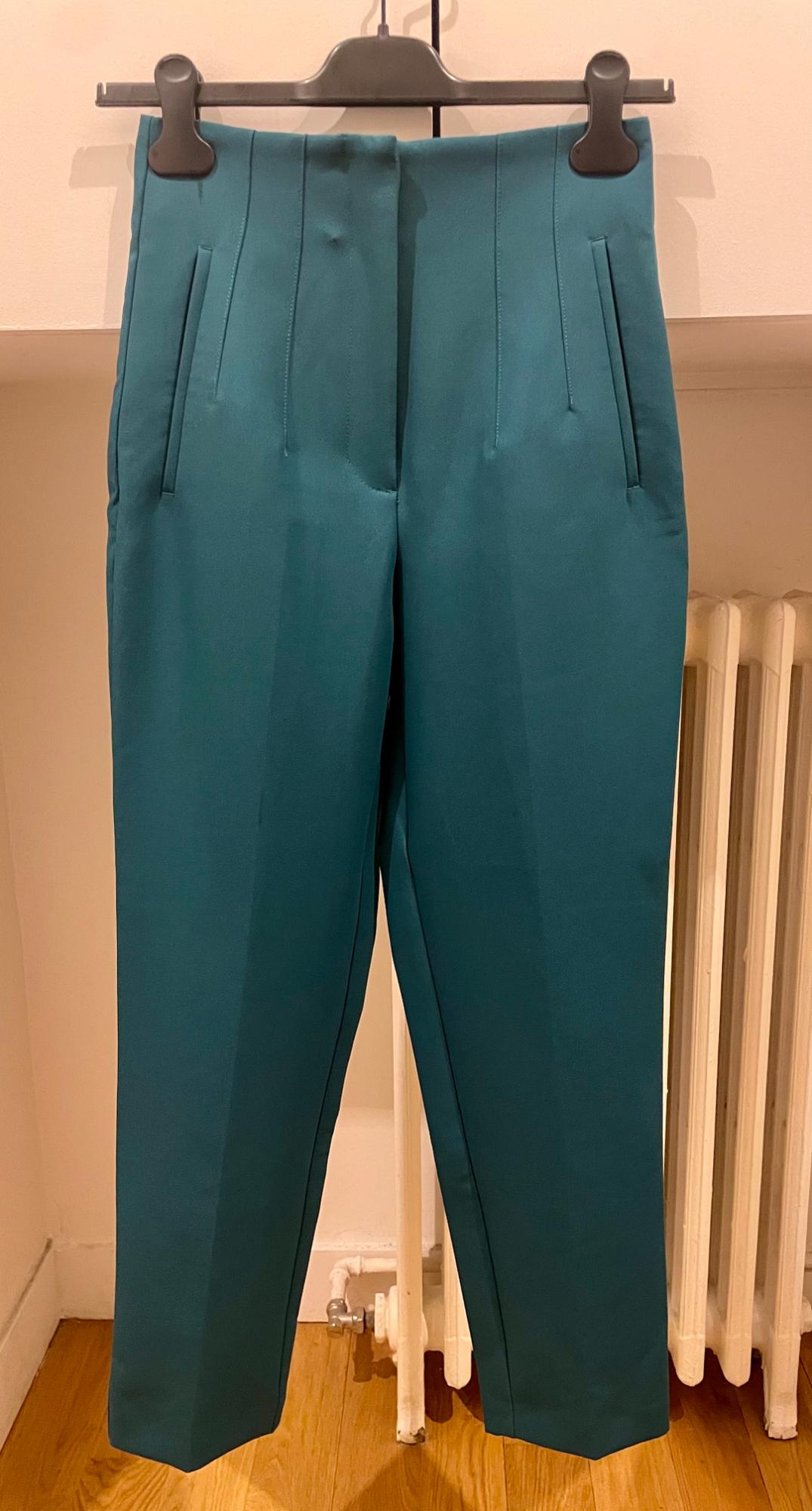 Teal trousers hanging on a black hanger against a white wall background and light wooden floor.