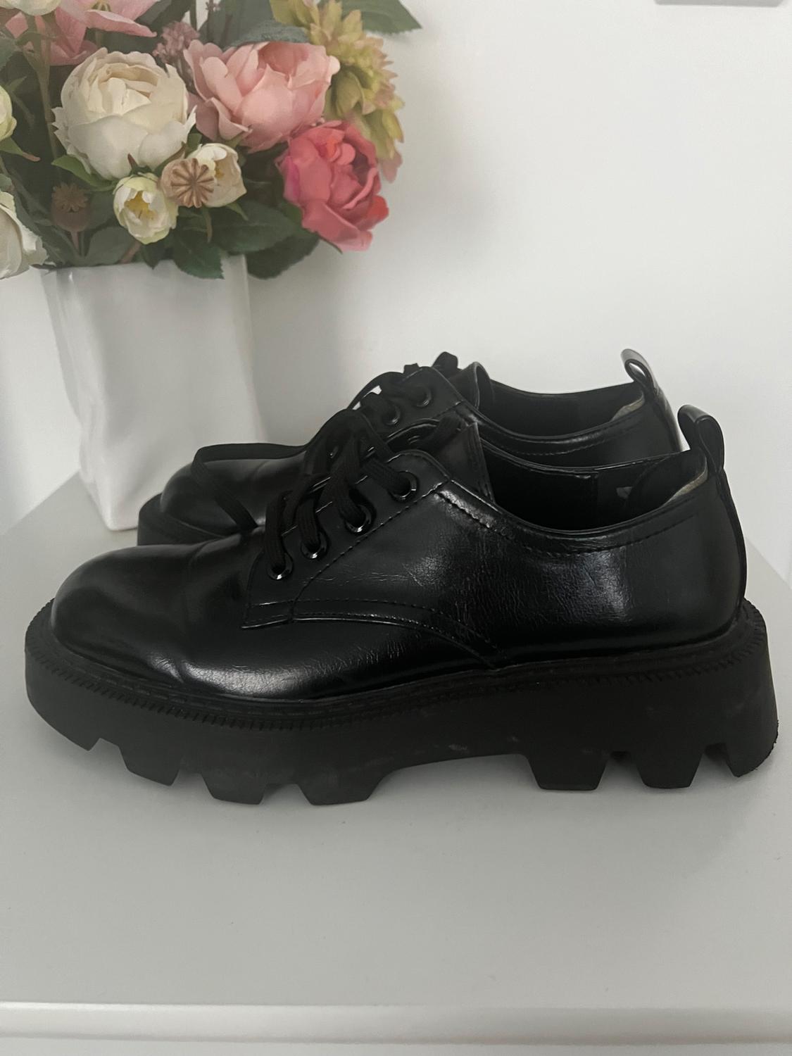 Black leather shoes with thick soles on a white table, next to a vase with colourful flowers.