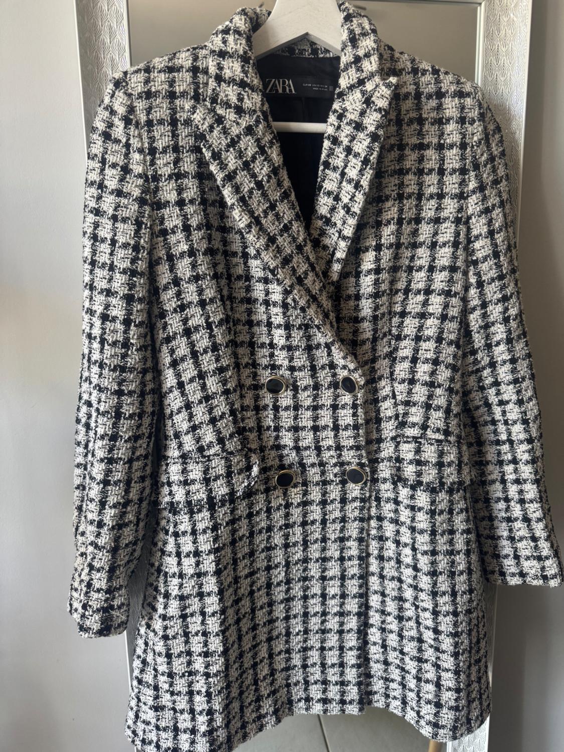 Black and white checked double-breasted coat on a hanger in front of a mirror. Four buttons and the label with the text ZARA stand out.
