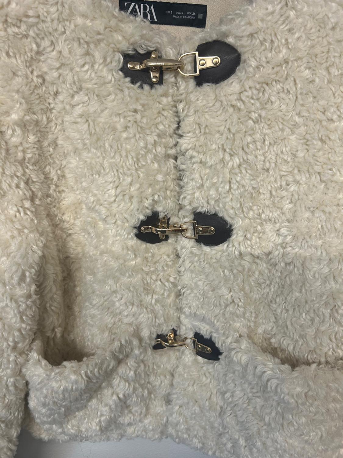Close-up of a textured cream jacket with gold fasteners and black patches. Text: ZARA EUR S USA S MEX 26 MADE IN CAMBODIA.