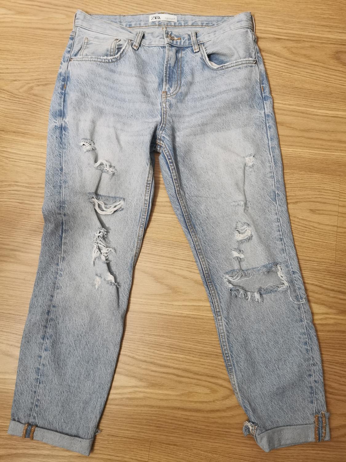 Light blue denim jeans with rips and turned-up hems, on a wooden background. The internal label reads ZARA.