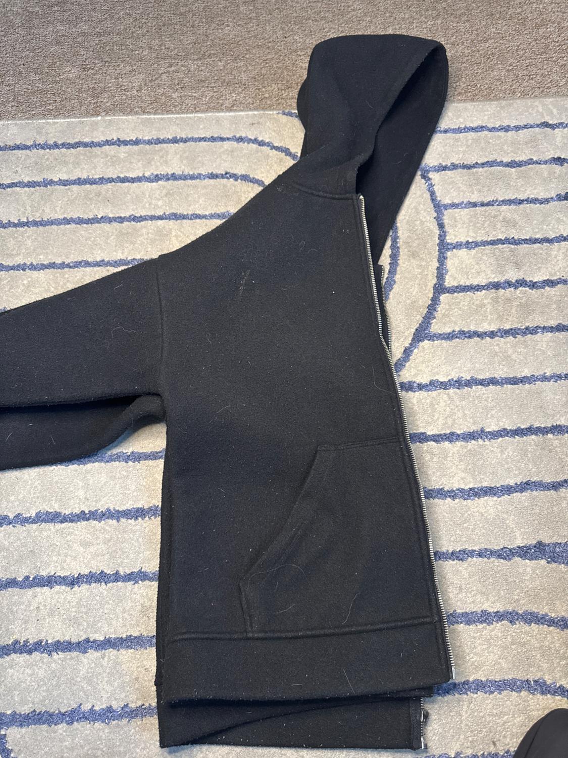Black hooded zip-up sweatshirt, folded on a grey rug with blue stripes. Relaxed, casual style composition.