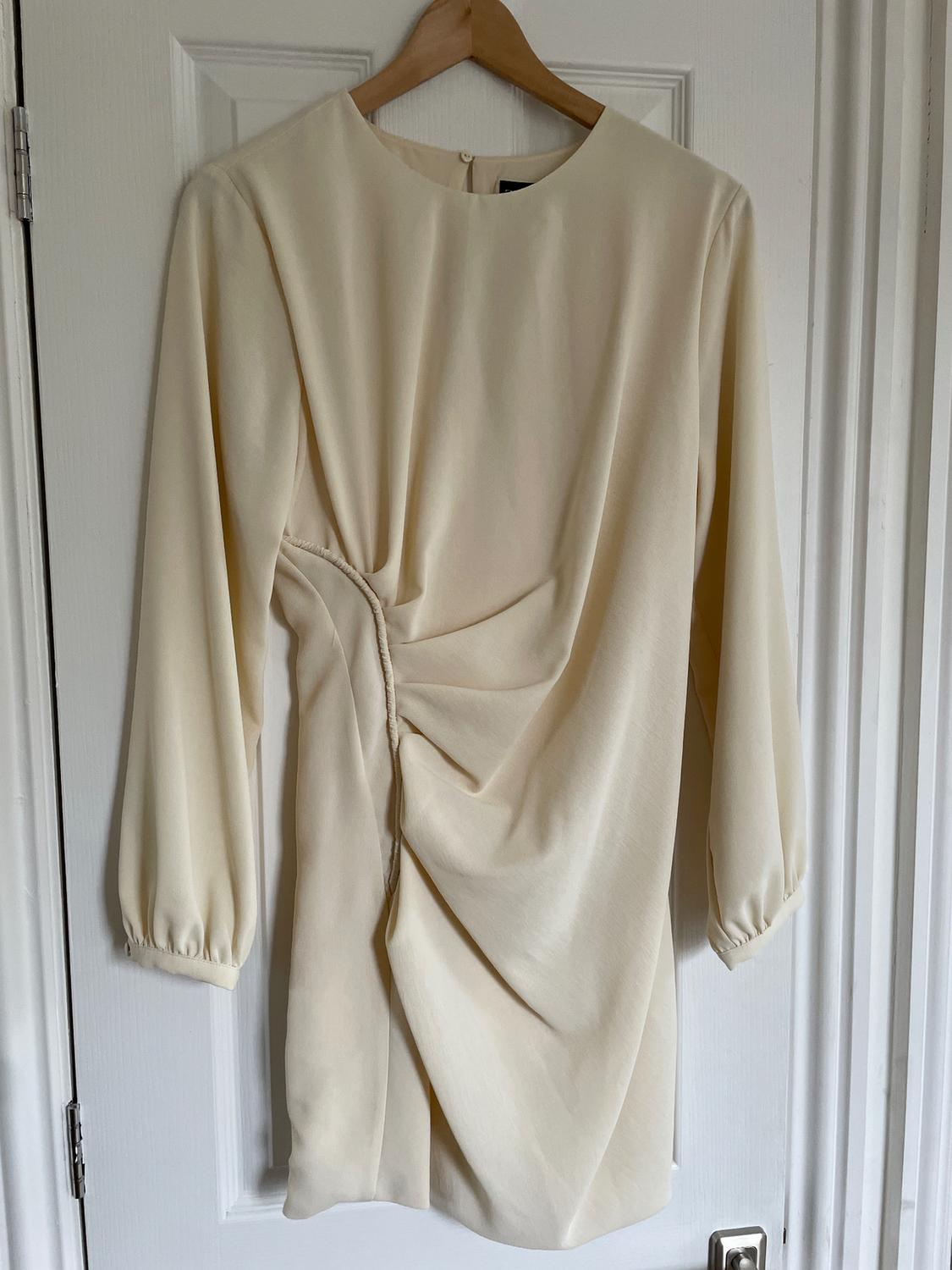 Long-sleeved beige dress with pleats at the side, hanging on a wooden hanger over a white door.