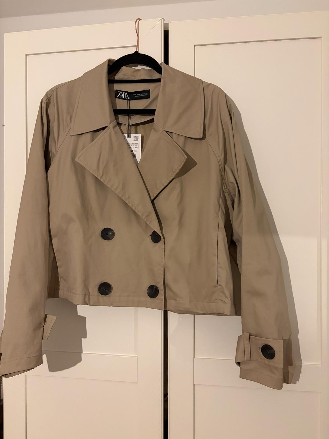 Beige short jacket with large black buttons, hanging on a hanger in front of a white wardrobe. Label visible on the collar.