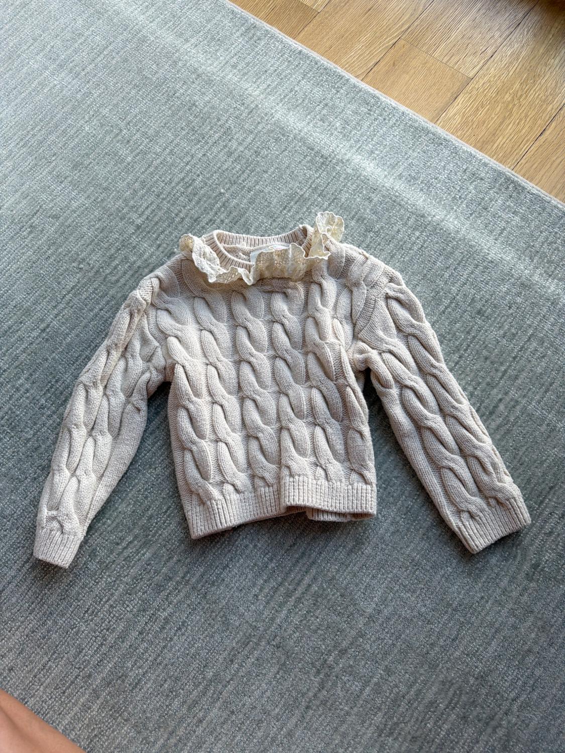 Chunky knit beige jumper with a frill collar, on a grey rug and light wooden floor.