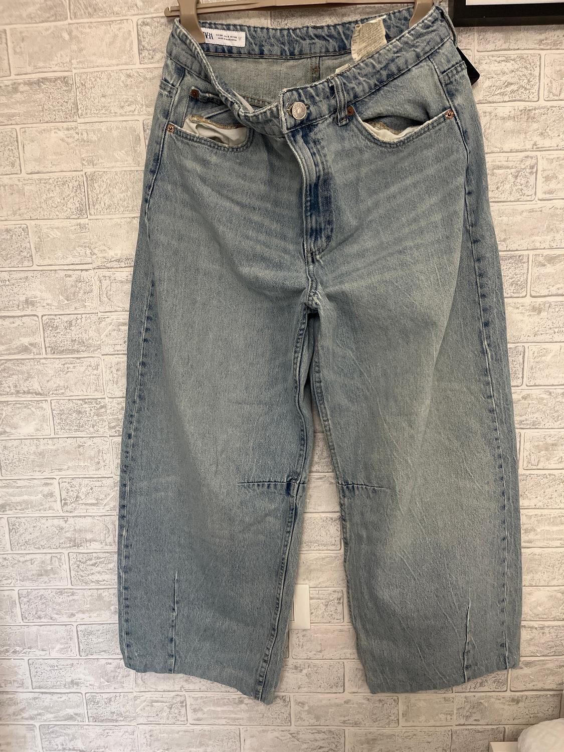 Light blue denim jeans hanging against a white brick wall. Visible pockets and waist label.