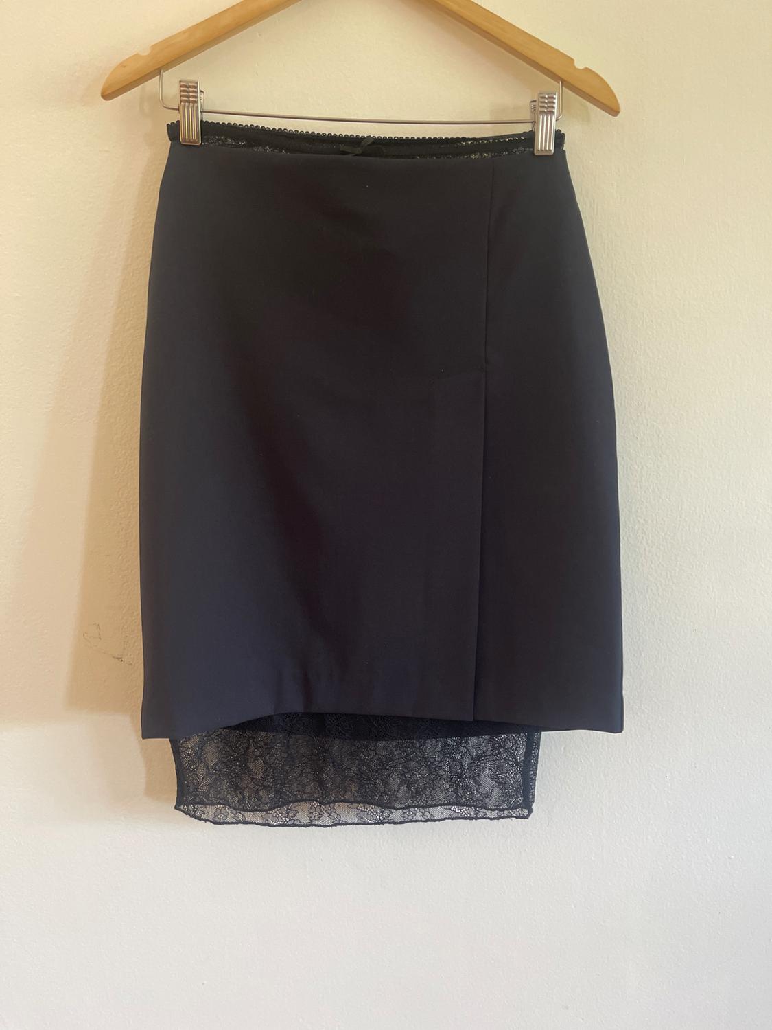 Black skirt on a wooden hanger against a white wall. A semi-sheer lace panel stands out, protruding from the hem.