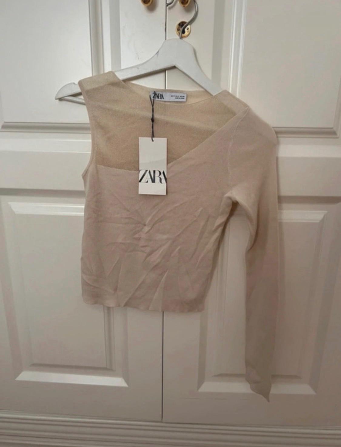 Beige asymmetric top with semi-transparent detail and one long sleeve, hanging on a white door. Label with the text ZARA.