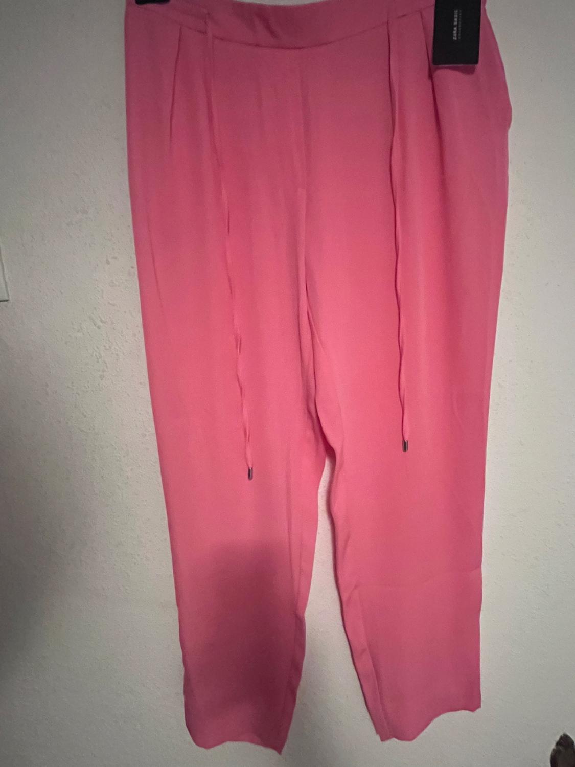 Pink trousers hanging on a hanger against a white wall. Elasticated waist with visible drawstrings.