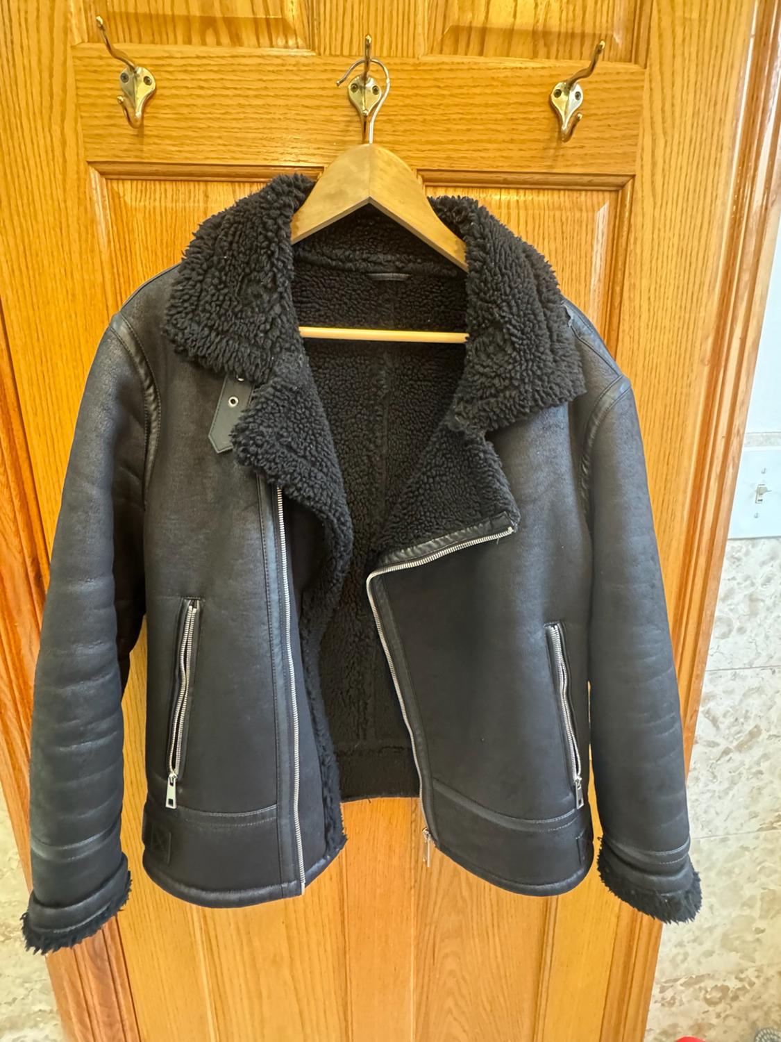 Black aviator-style jacket with faux fur lining and zips, hanging on a hanger over a wooden door in a casual setting.