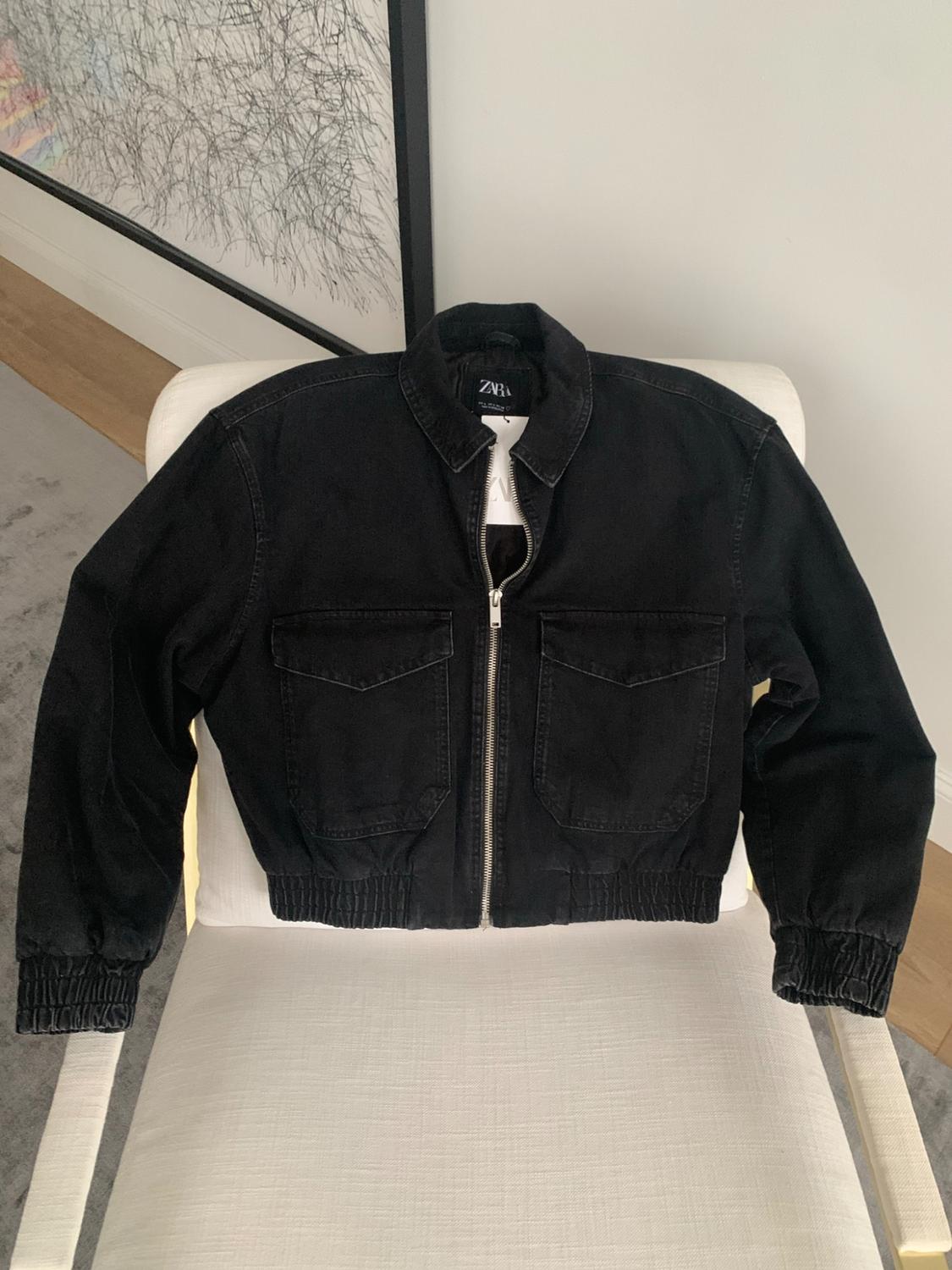 Black zip-up jacket with flap pockets and elastic hems, draped over a white chair. Interior label with the text Zara.