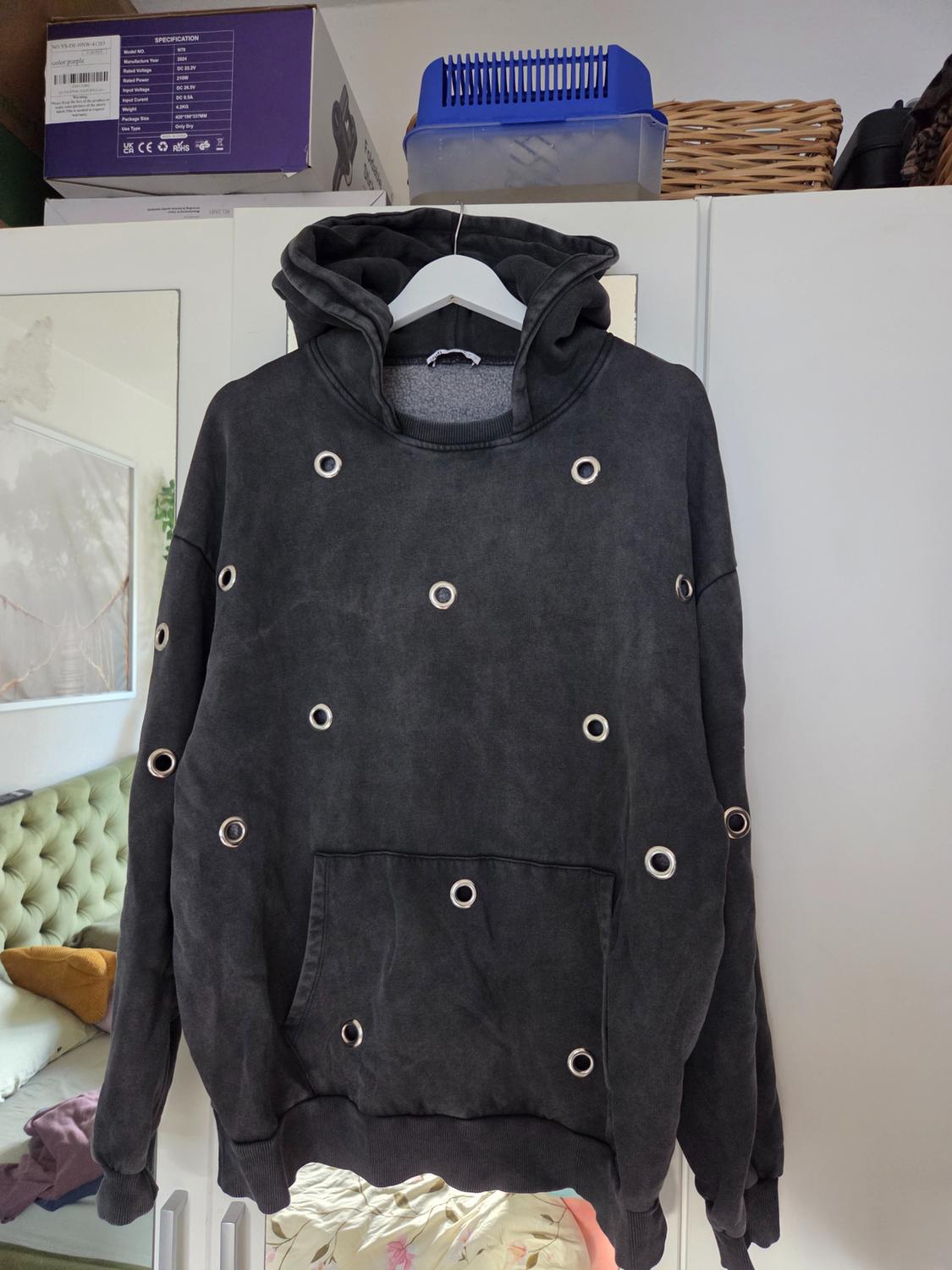 Worn dark grey hooded sweatshirt with metal rings hanging in a room. Text visible on a box: SPECIFICATION.